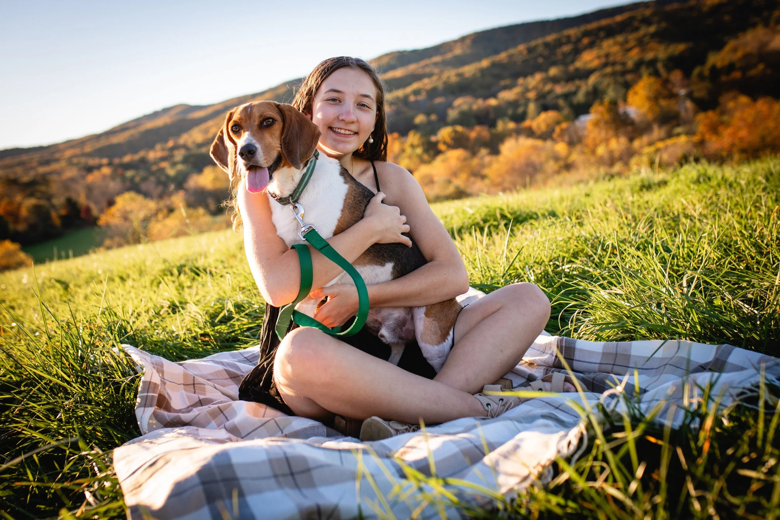 A young girl sitting on a checkered blanket in a grassy field, hugging a beagle dog, with autumn-colored trees and mountains in the background.