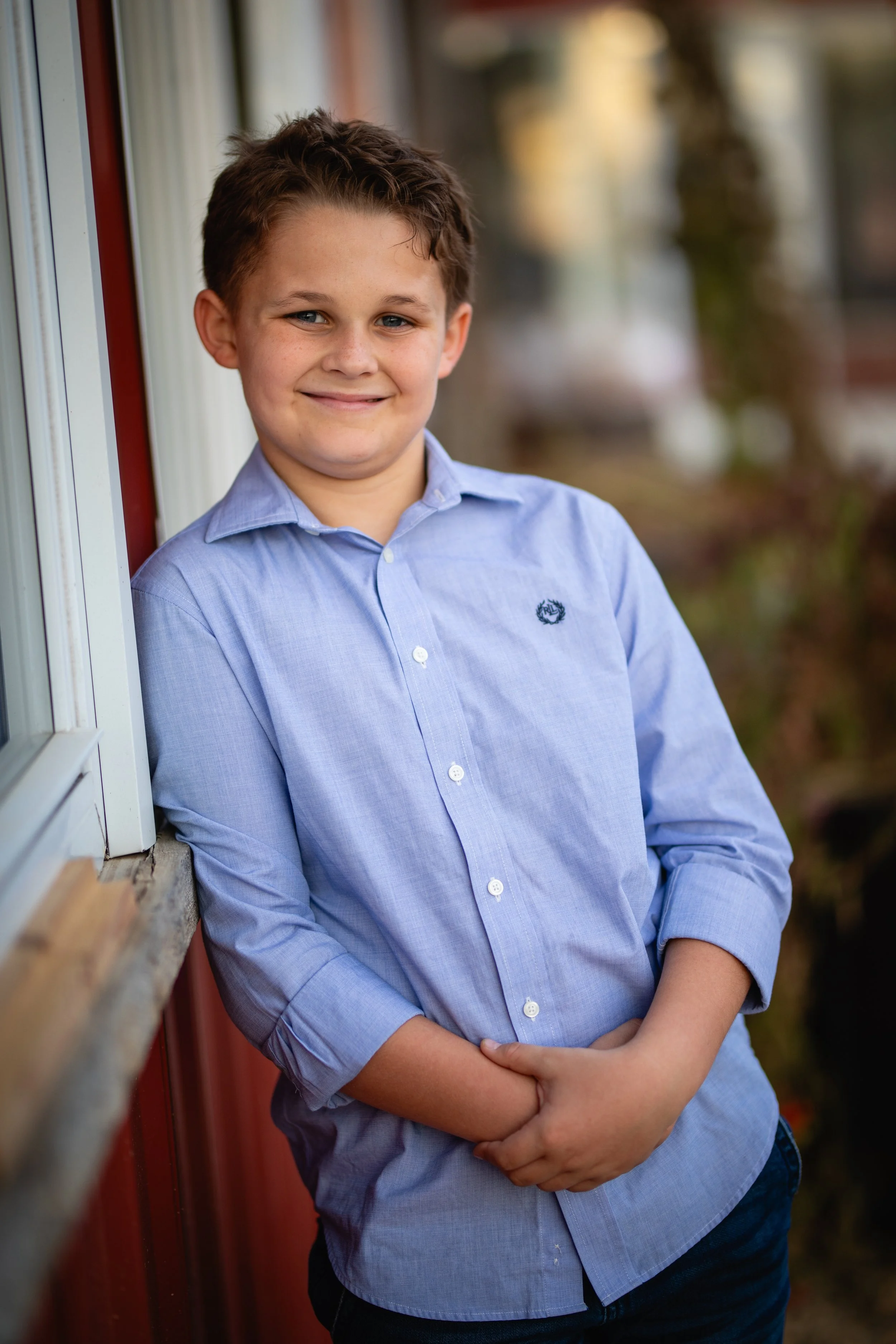 A young boy with short brown hair, smiling, wearing a light blue button-up shirt and dark pants, leaning against a window with a blurred outdoor background.