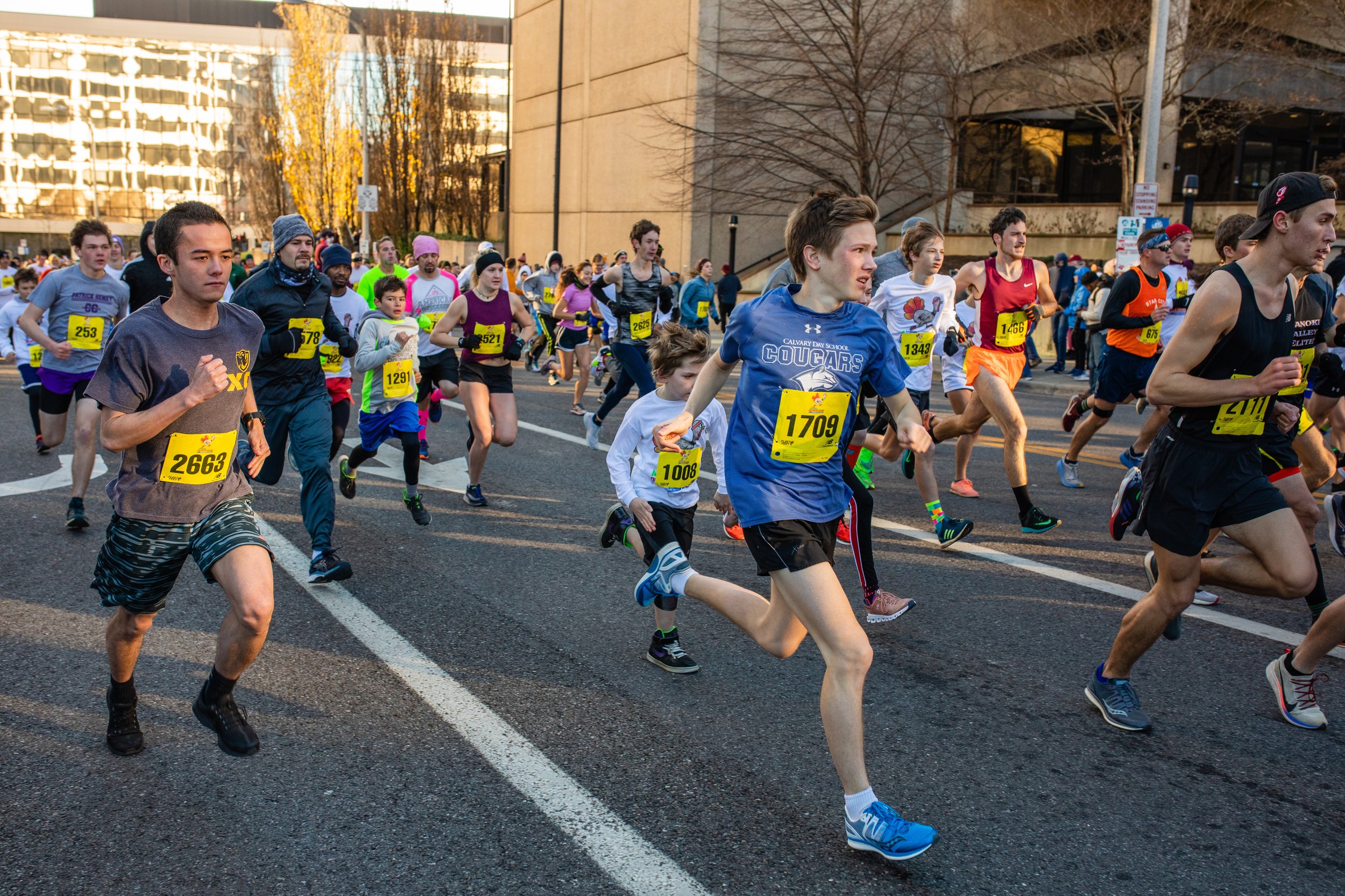 A large group of runners participating in a marathon on a city street during daylight, with some runners wearing race bibs and athletic clothing.