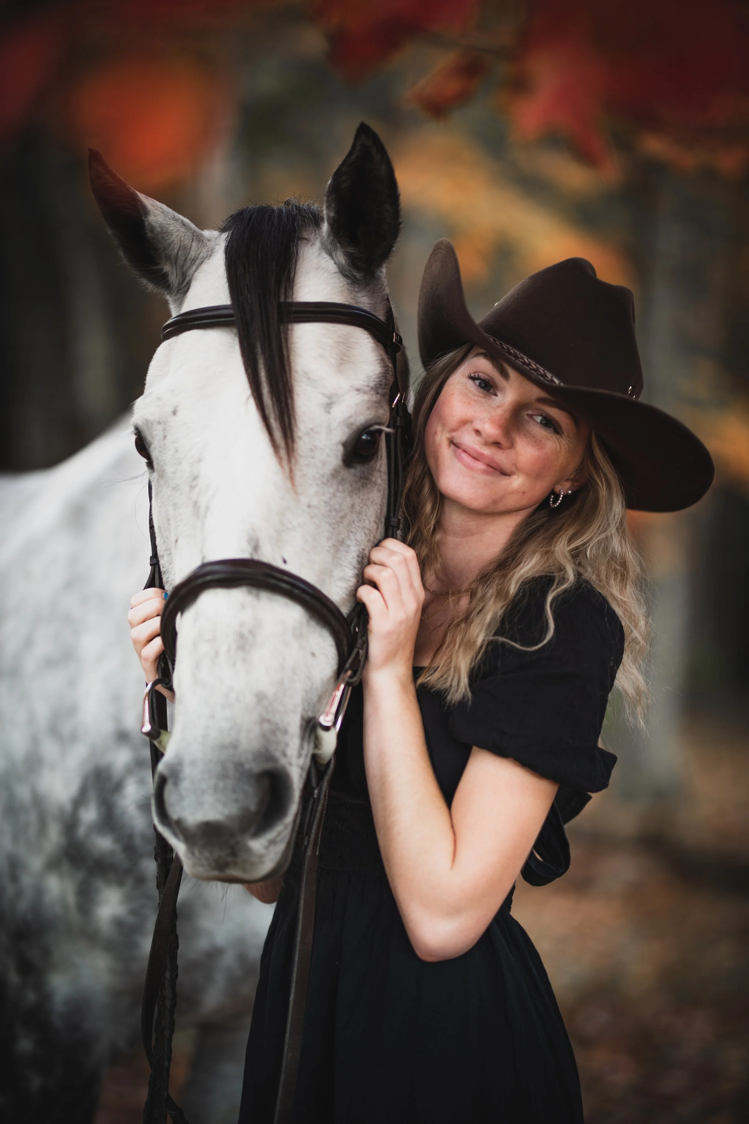A young woman with wavy blonde hair wearing a black dress and a wide-brimmed brown hat, standing close to a white horse with black markings on its face in an outdoor setting with autumn-colored leaves in the background.