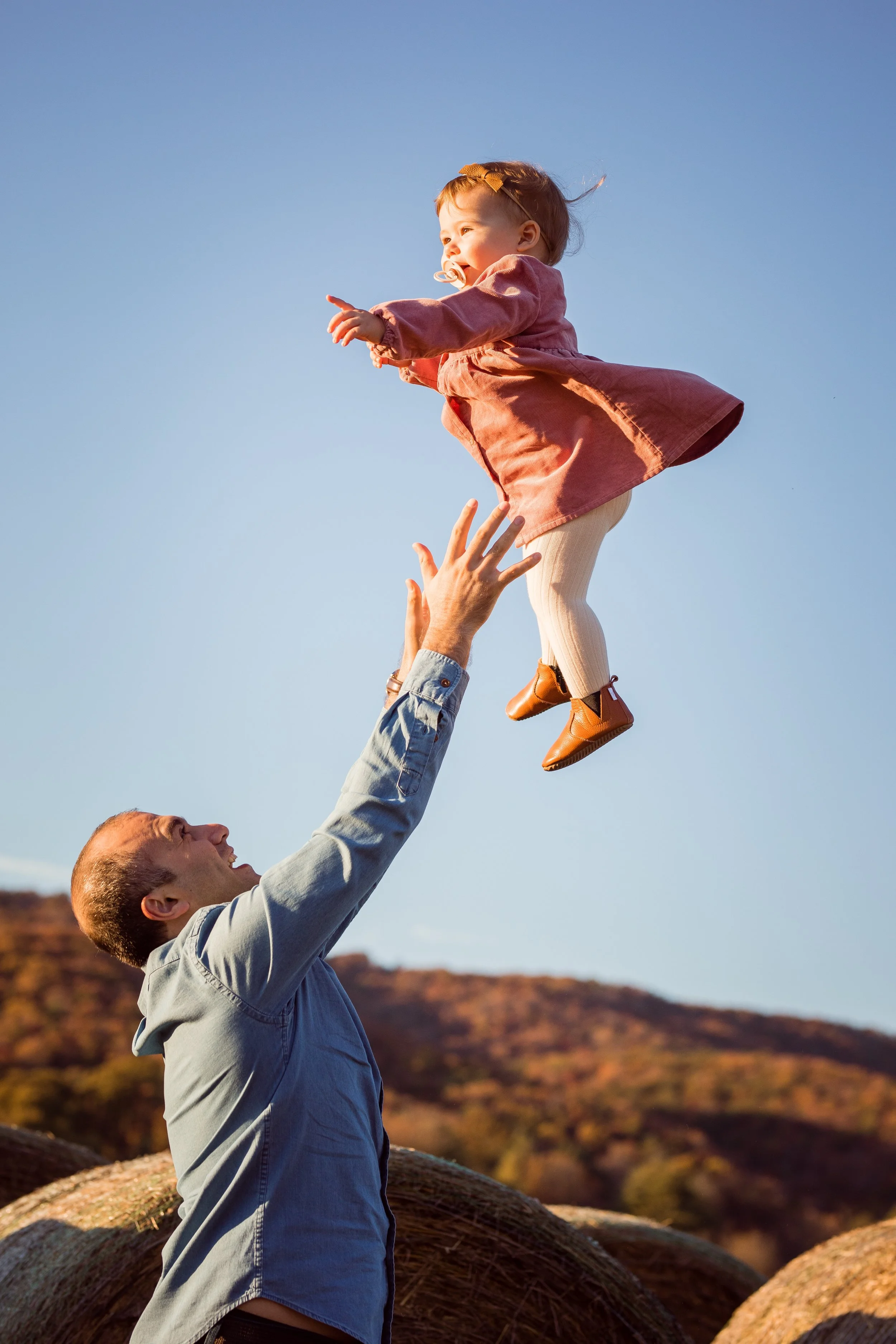 A man lifting a young girl into the air outdoors at sunset with hay bales and a hillside in the background.