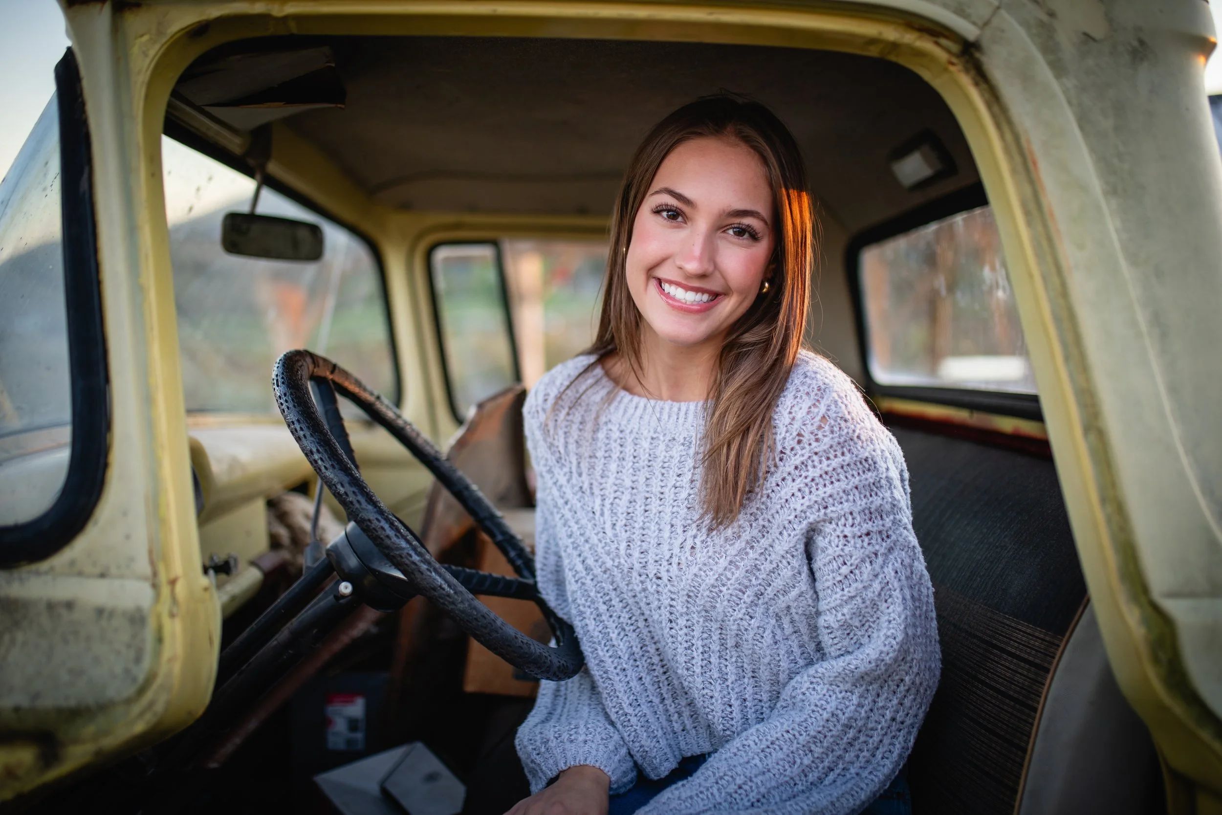 A young woman with long brown hair and a bright smile sitting inside an old yellow truck, wearing a cozy gray sweater.