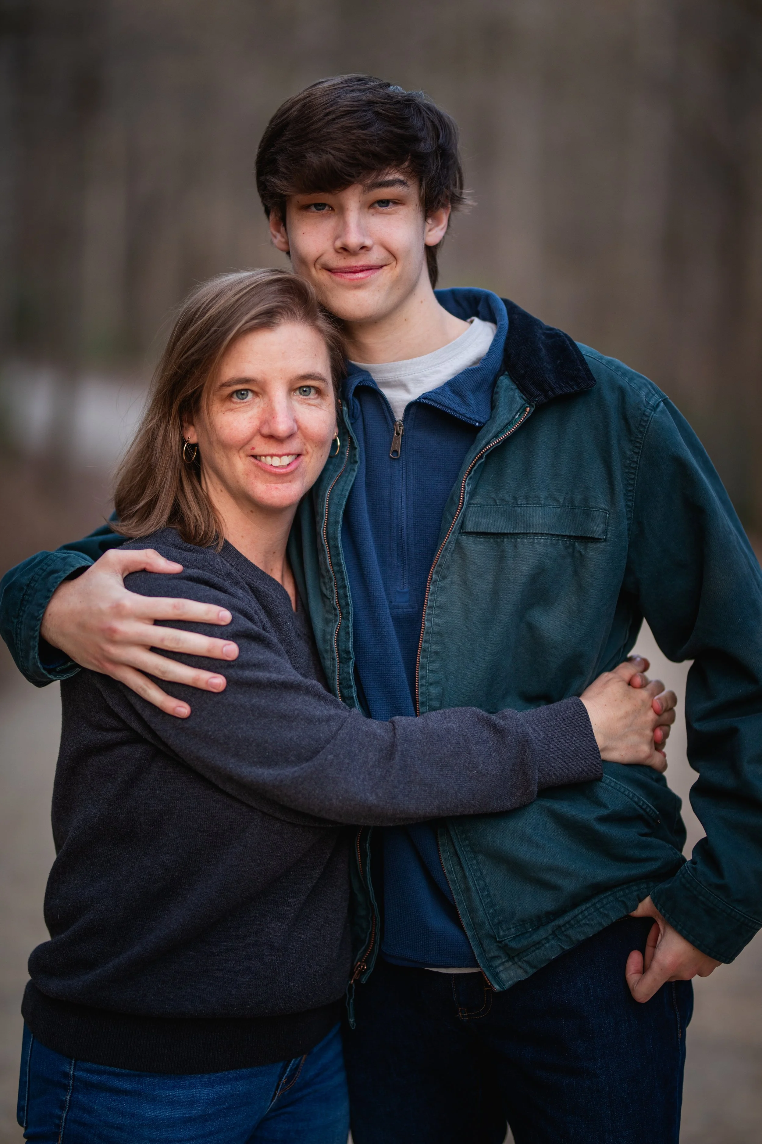 A woman and a teenage boy standing outdoors, embracing each other, with trees in the background.