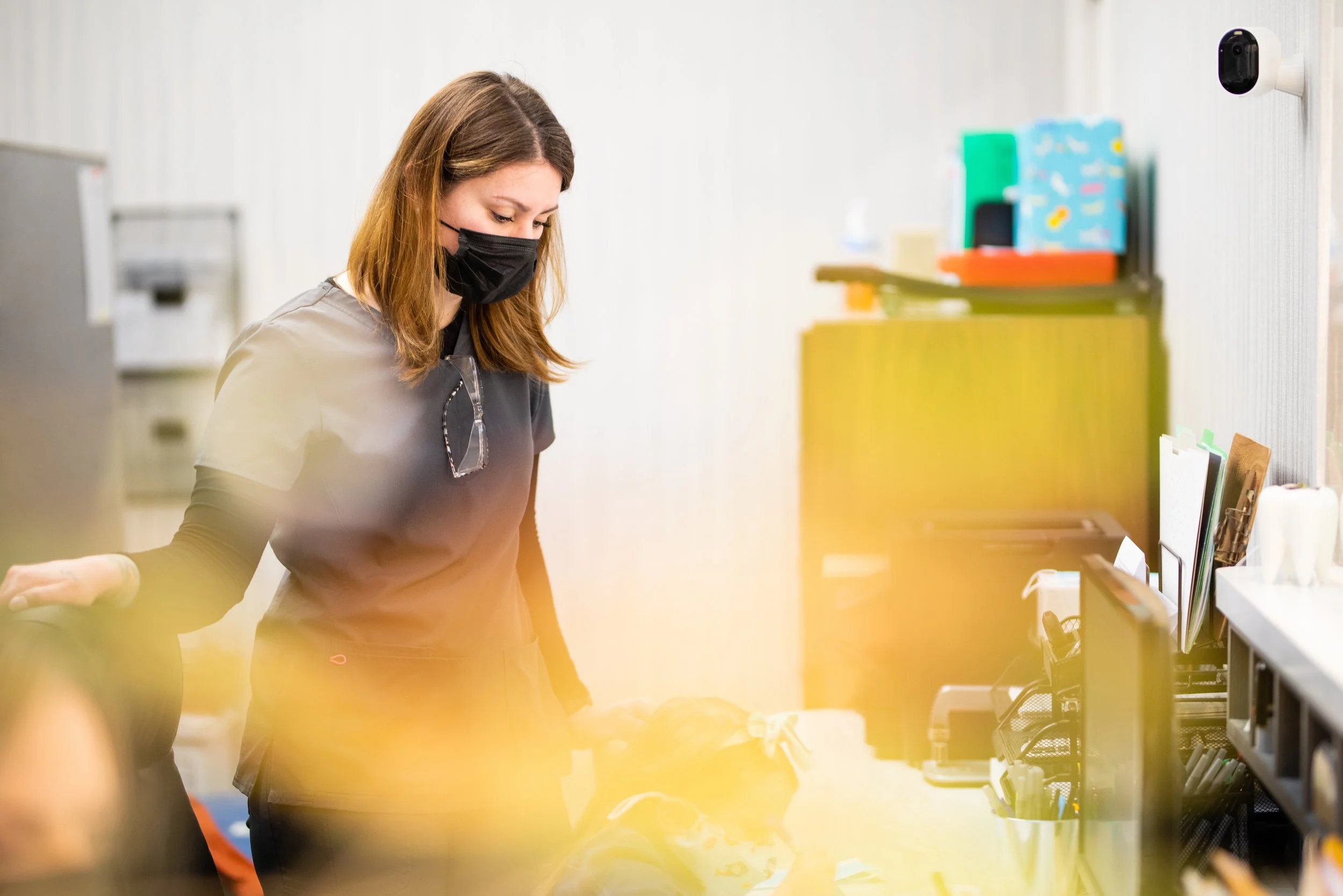 A woman wearing a black face mask and glasses hanging from her shirt collar, looking down at a desk with office supplies, in an office environment.