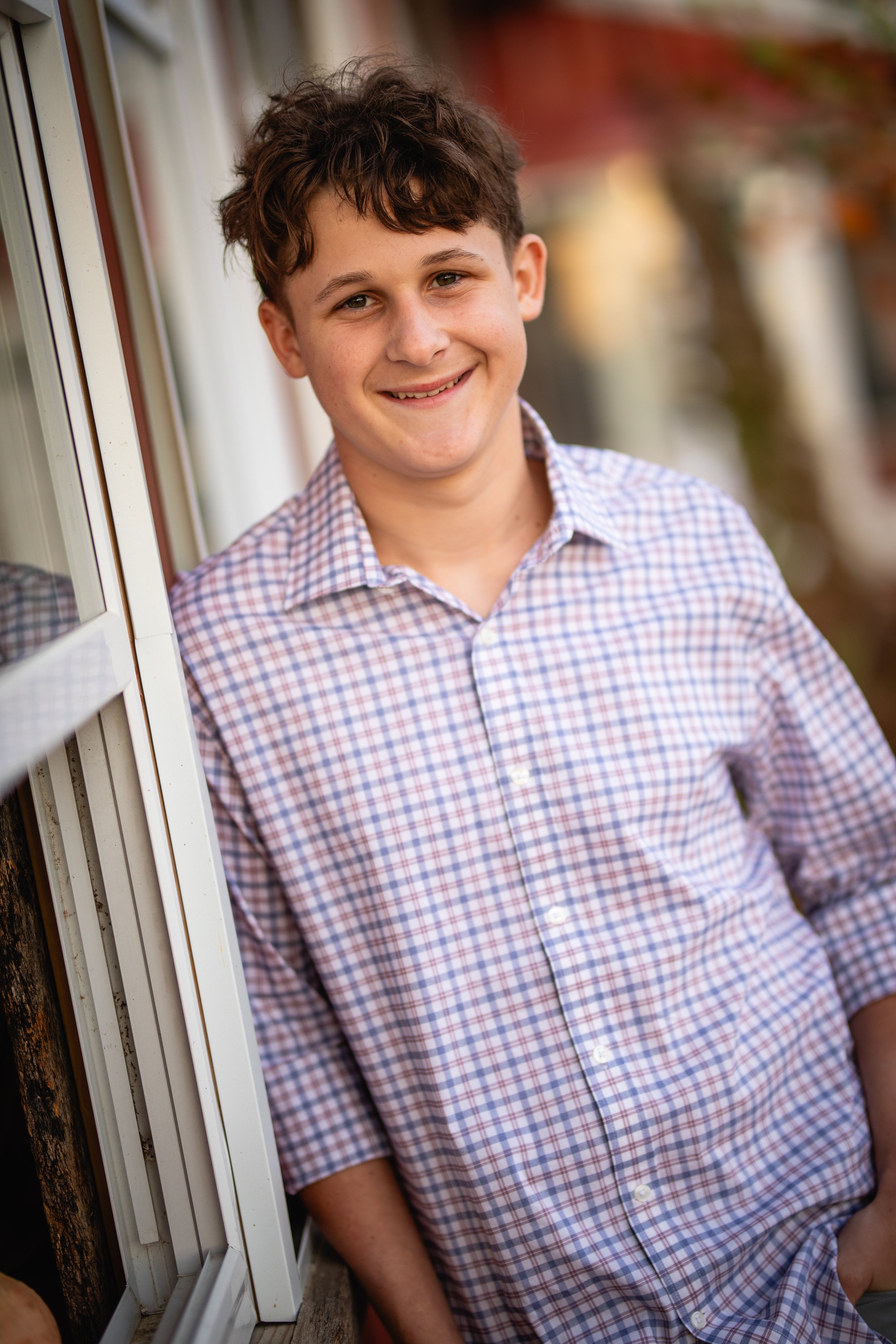 Young boy with brown curly hair smiling, wearing a checkered shirt, leaning against a window outside.