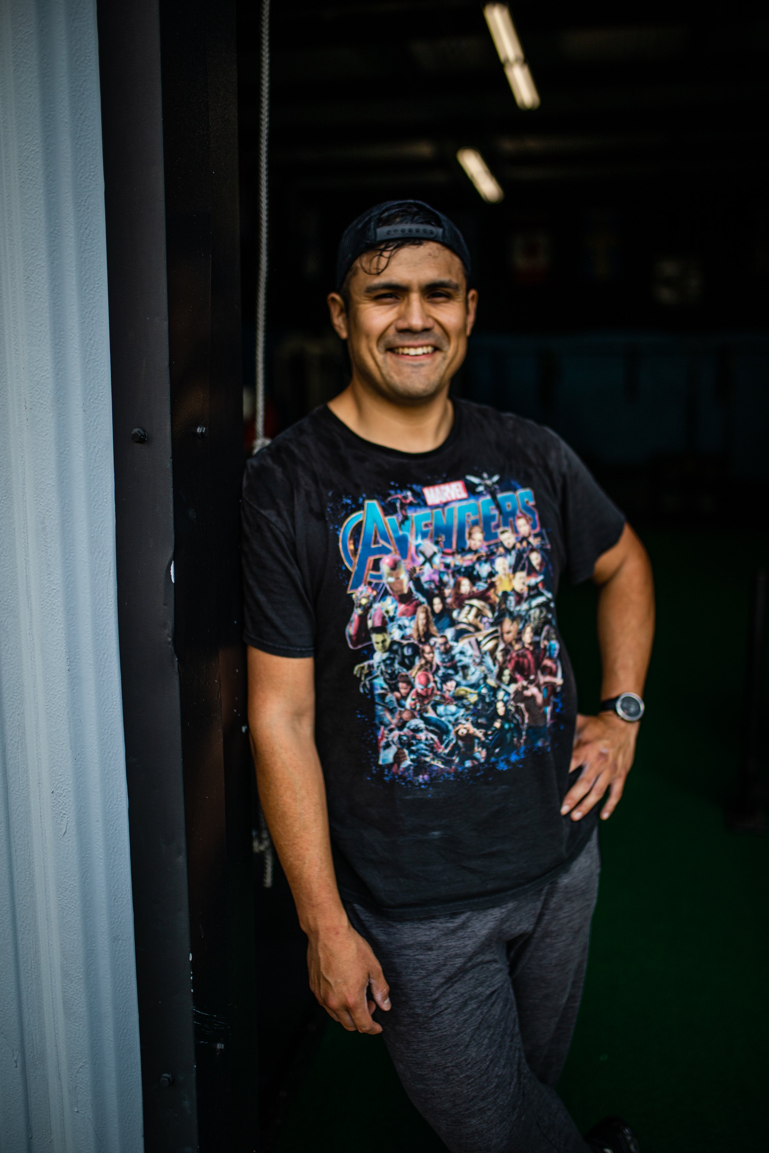 A man smiling while leaning against a wall inside a gym or training facility, wearing a black Avengers t-shirt, grey athletic pants, a cap, and a smartwatch.