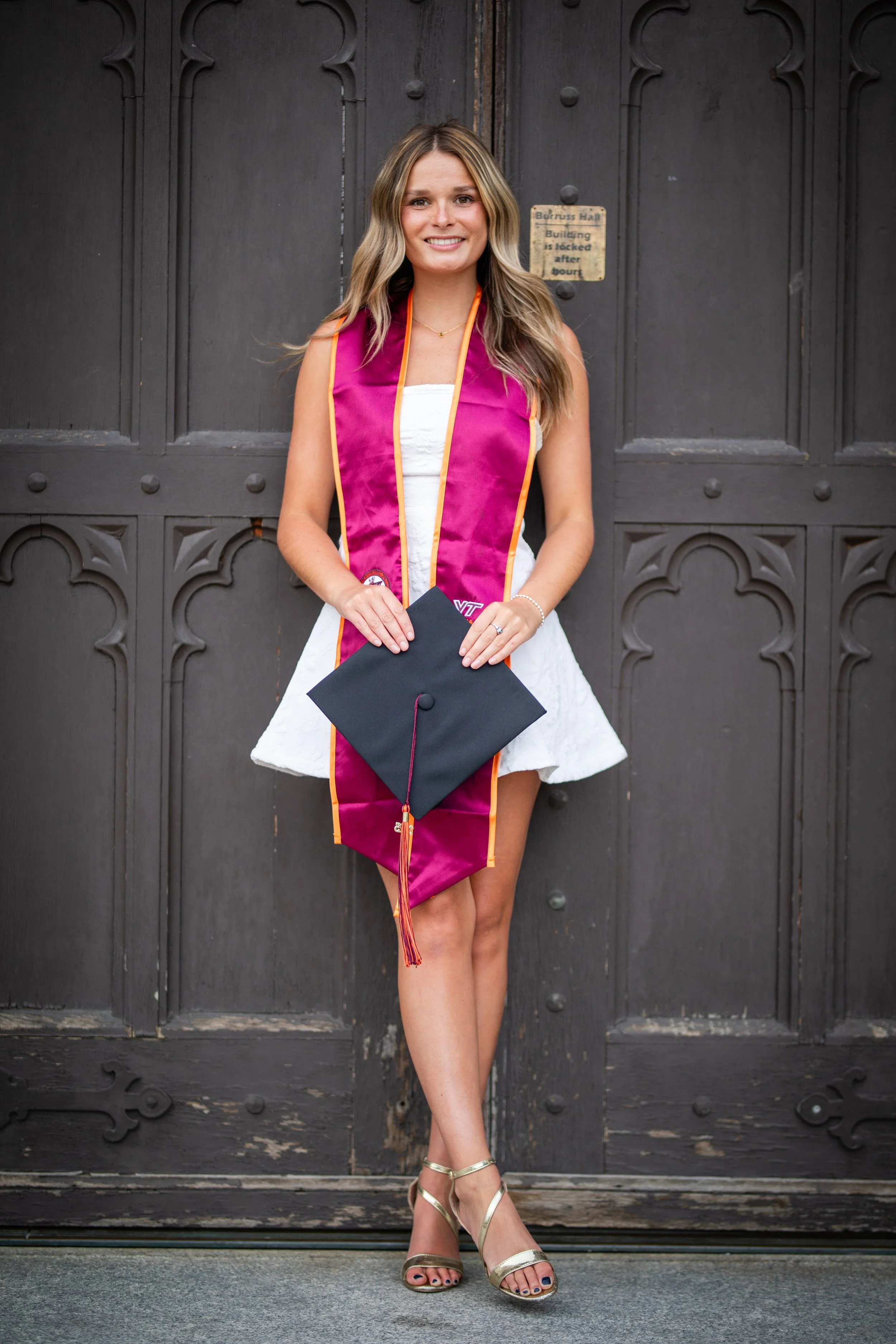 A young woman dressed in a white dress and gold heels holding a graduation cap, wearing a pink graduation stole, standing in front of a large black wooden door.