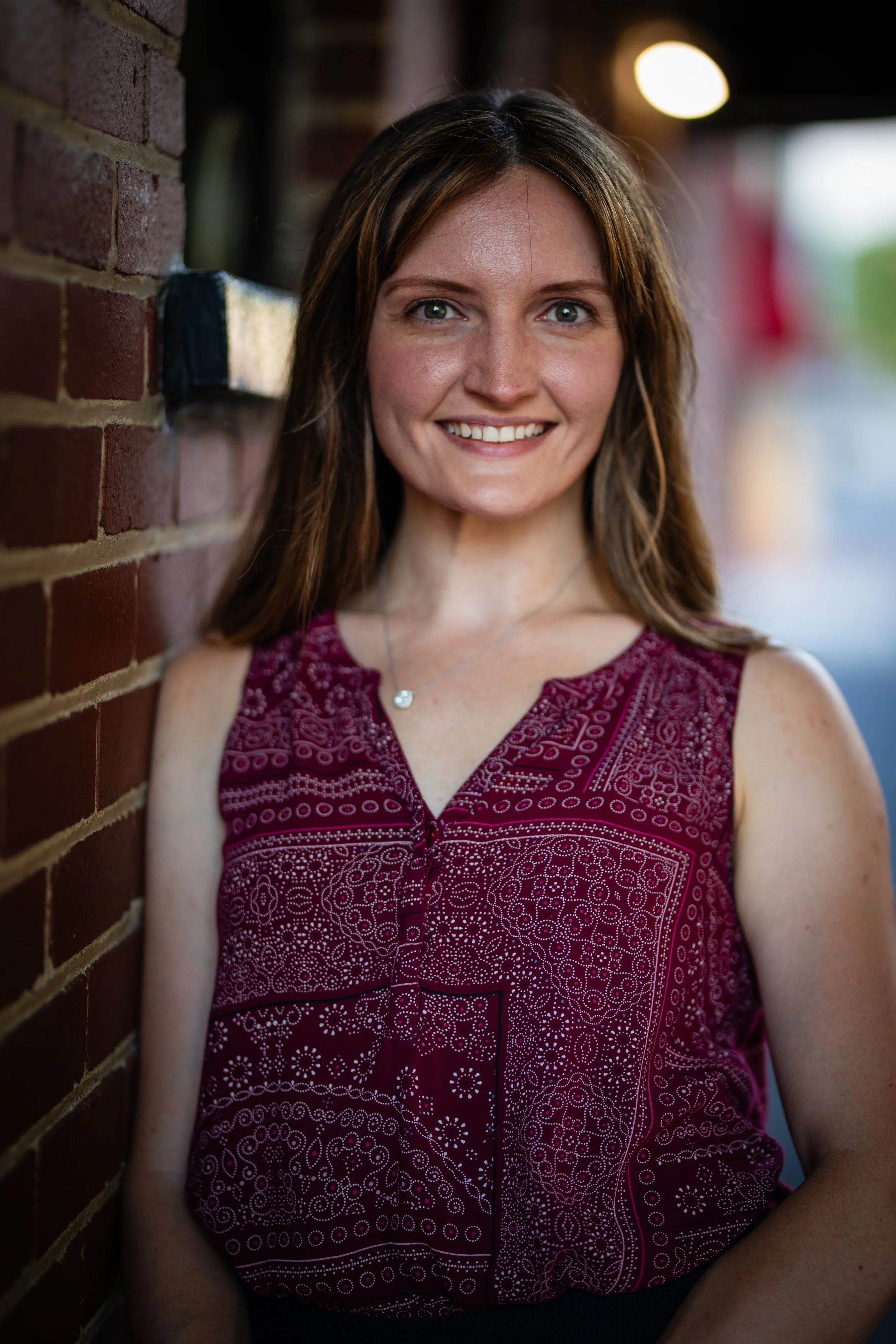 A woman with long brown hair and a wide smile, wearing a sleeveless red patterned top, standing against a brick wall with a blurred background.