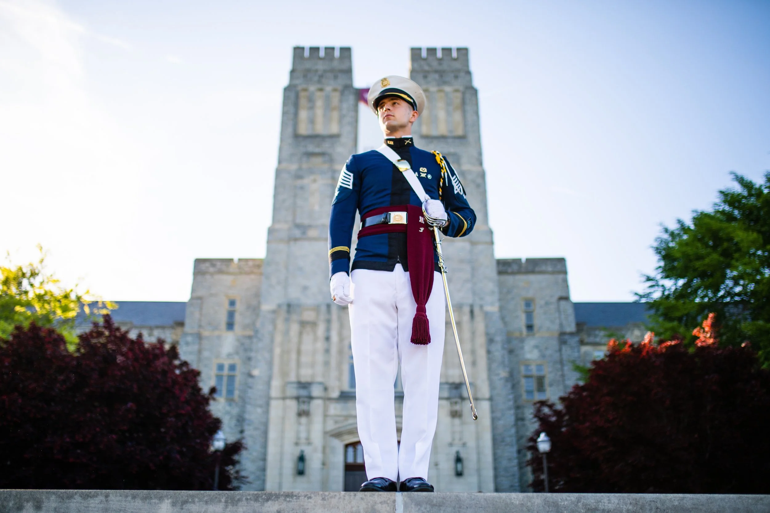 A young military cadet in dress uniform standing at attention in front of a stone building with two towers, with trees on either side.