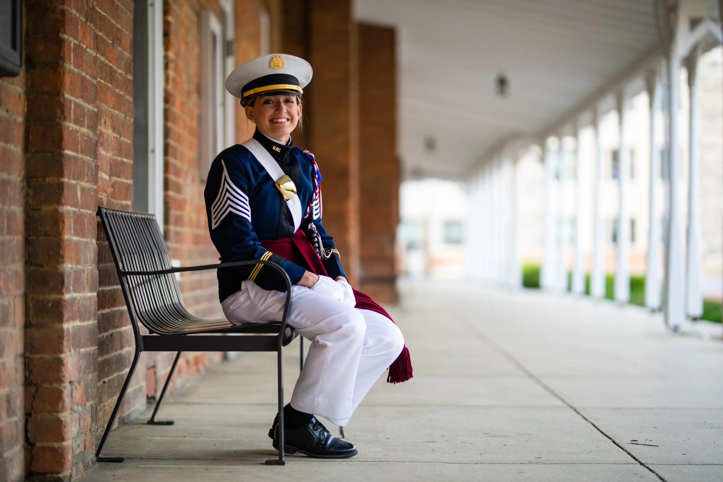 A young woman in a military uniform sitting on a bench outside a building with brick walls and white columns, smiling at the camera.