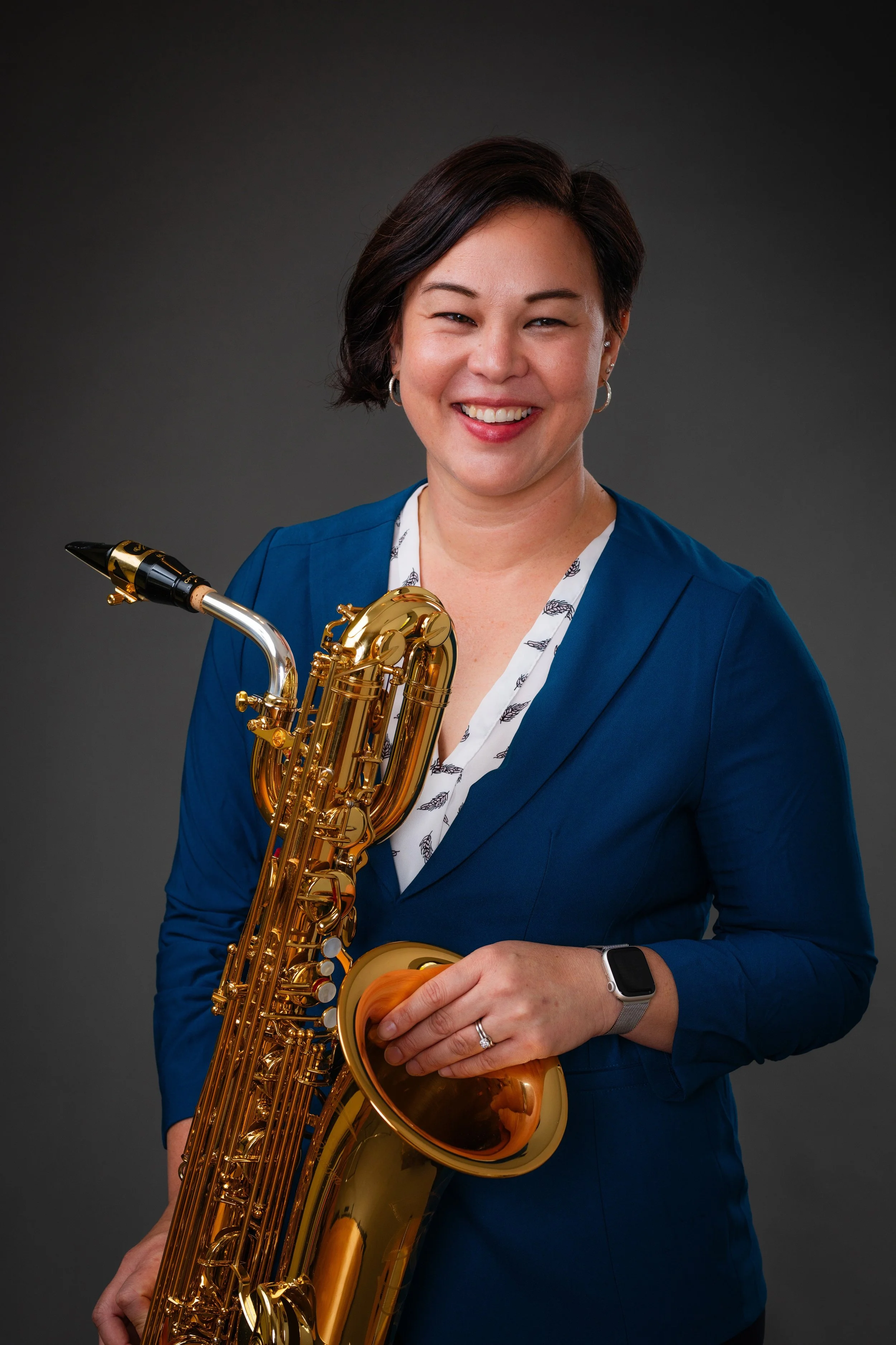 A woman with short dark hair smiling while holding a gold saxophone, wearing a blue blazer and a smartwatch on her left wrist, posed against a gray background.