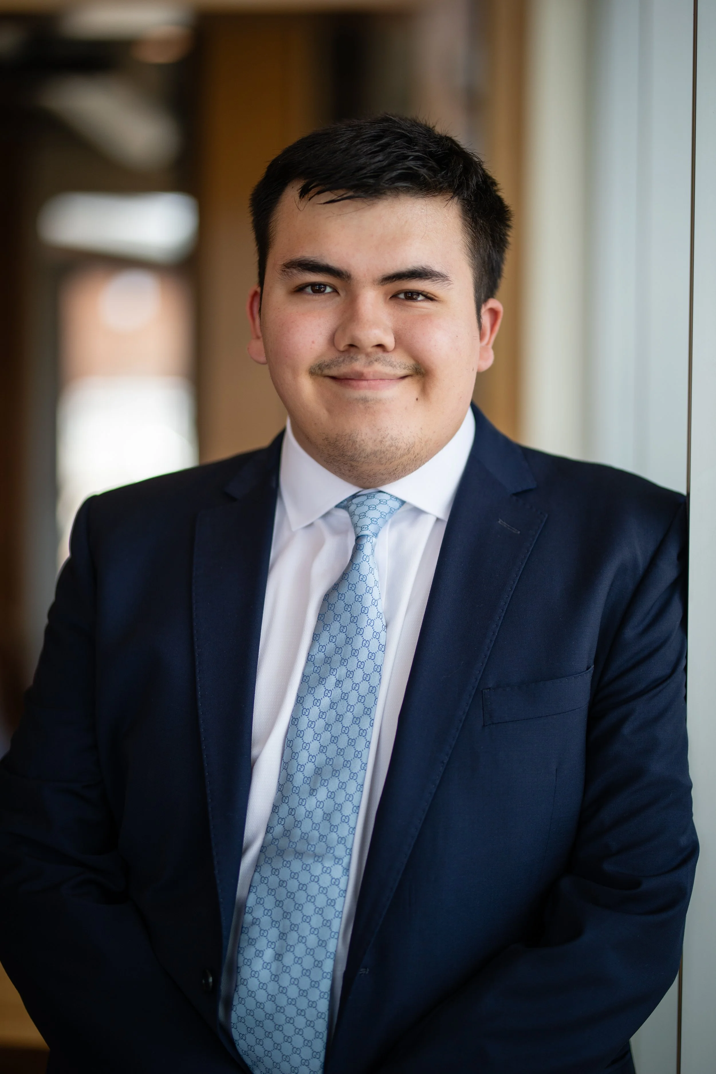 A young man in a navy blue suit, white shirt, and light blue patterned tie standing indoors with a neutral smile, natural light coming from the window on the right.