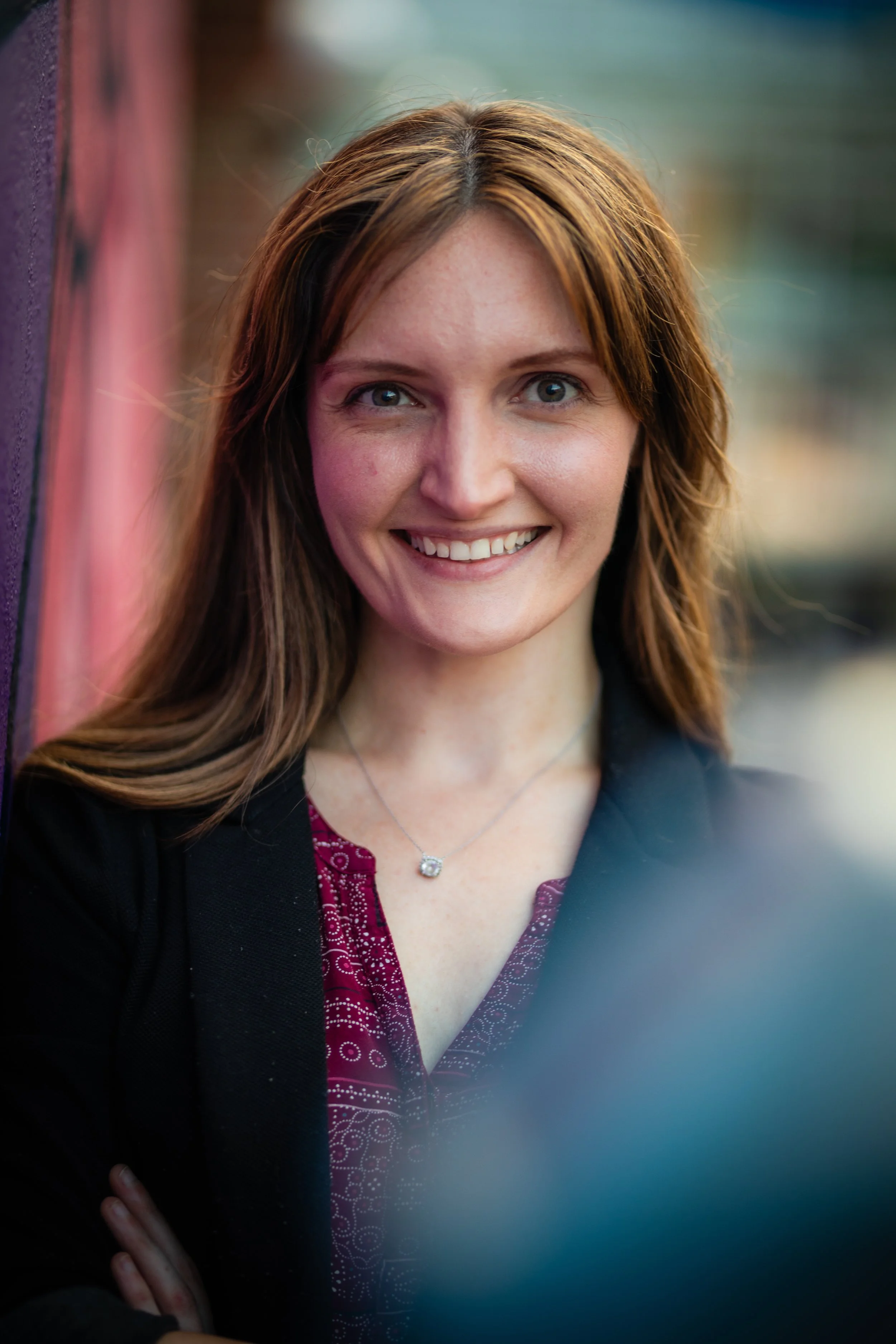 Close-up of a smiling woman with red hair, wearing a black jacket and a burgundy patterned shirt, standing outdoors near a colorful wall.