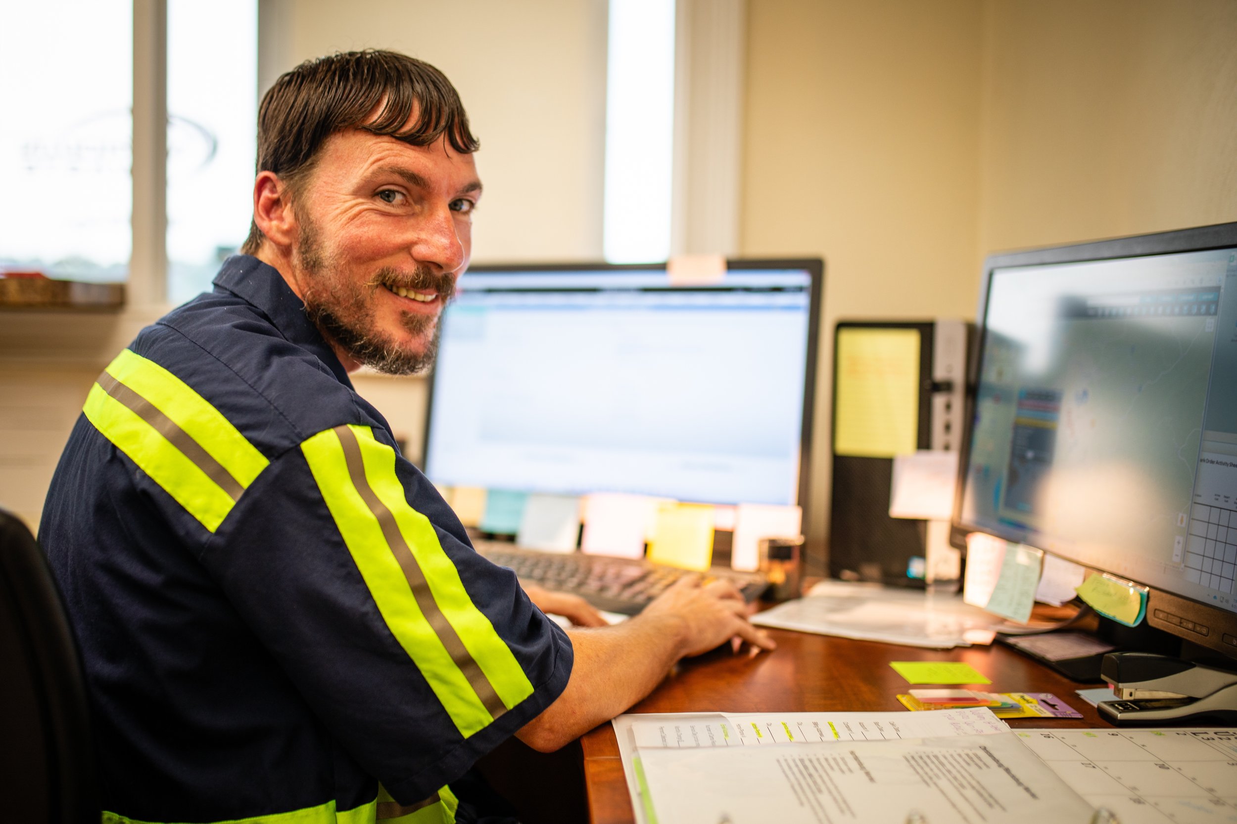 A man in a dark blue uniform with reflective yellow stripes sitting at a desk with two computer monitors, smiling at the camera.