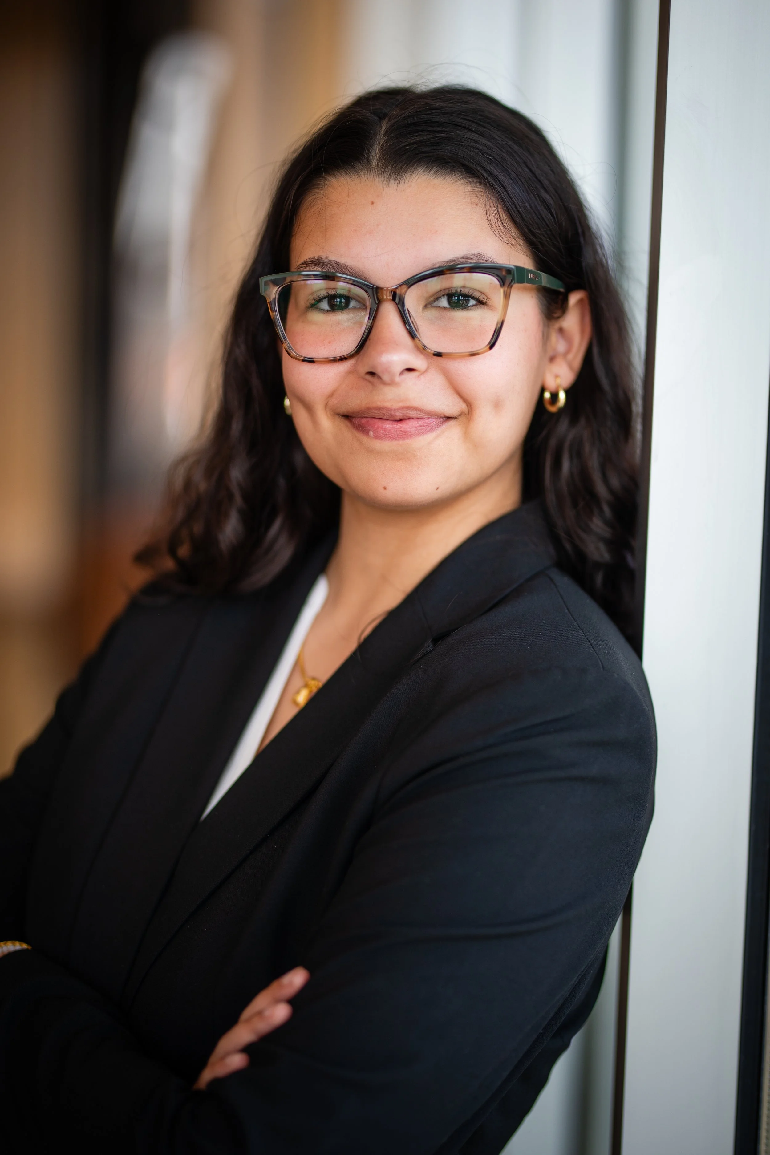 A confident woman with dark brown hair, glasses, gold earrings, and a heart-shaped necklace, wearing a black blazer, standing with her arms crossed near a window in an indoor setting.