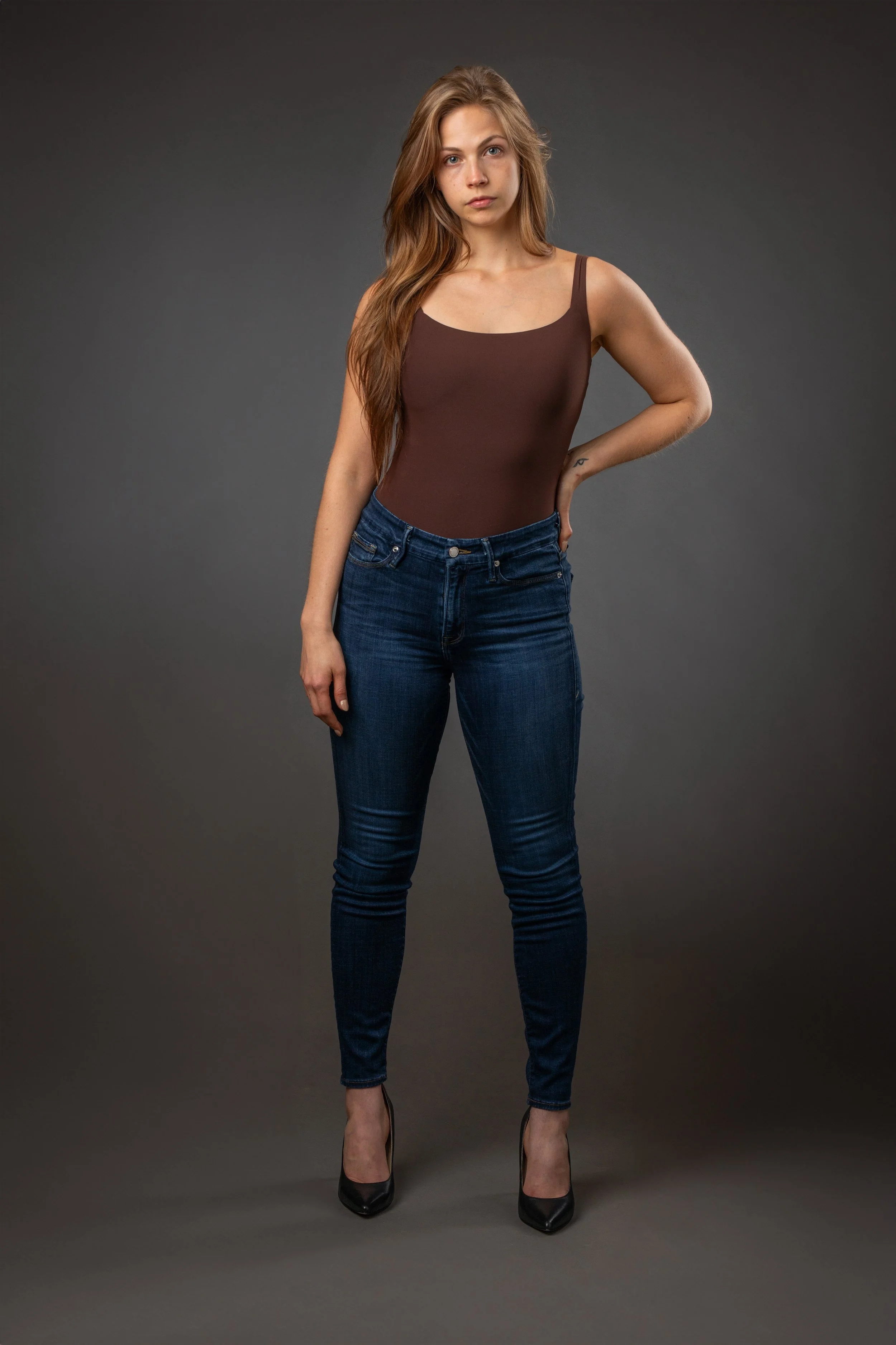 A young woman with long, wavy brown hair standing against a gray background, wearing a brown fitted tank top, dark blue jeans, and black high heels, looking at the camera with a neutral expression.