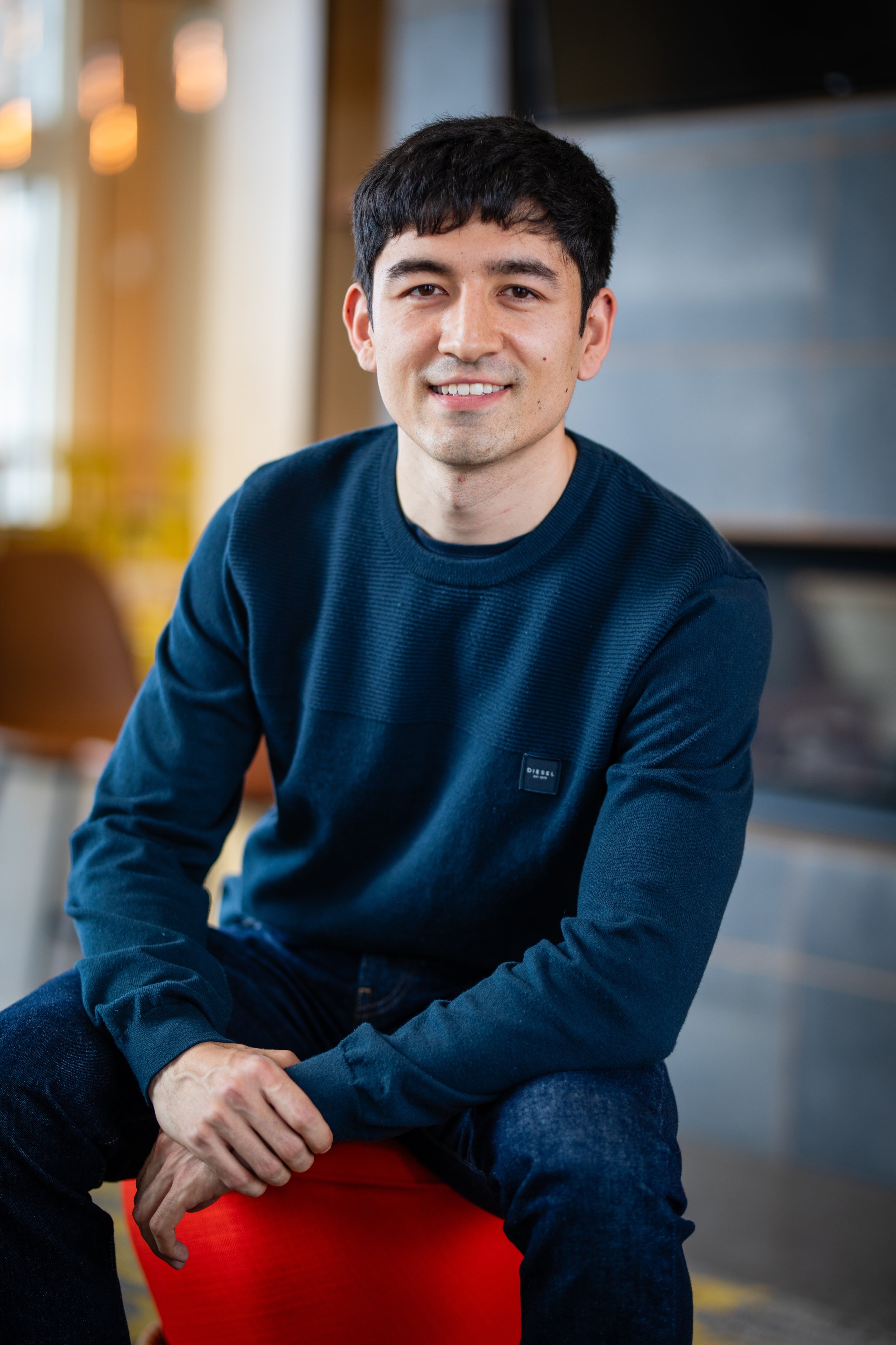 A young man with dark hair, wearing a navy blue sweater, is sitting on a red stool in a modern indoor space, smiling at the camera.