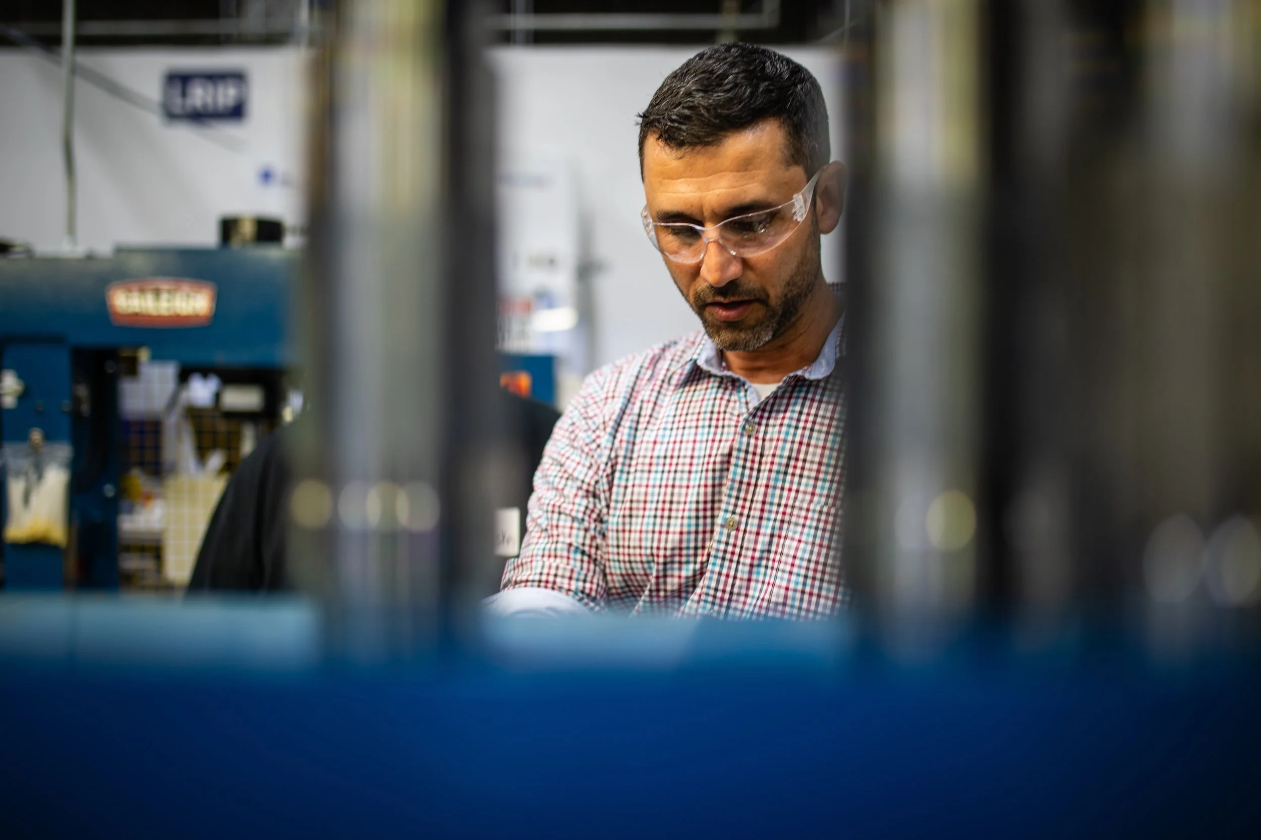 A man wearing safety glasses and a checkered shirt working in a lab or workshop, surrounded by equipment and tools.