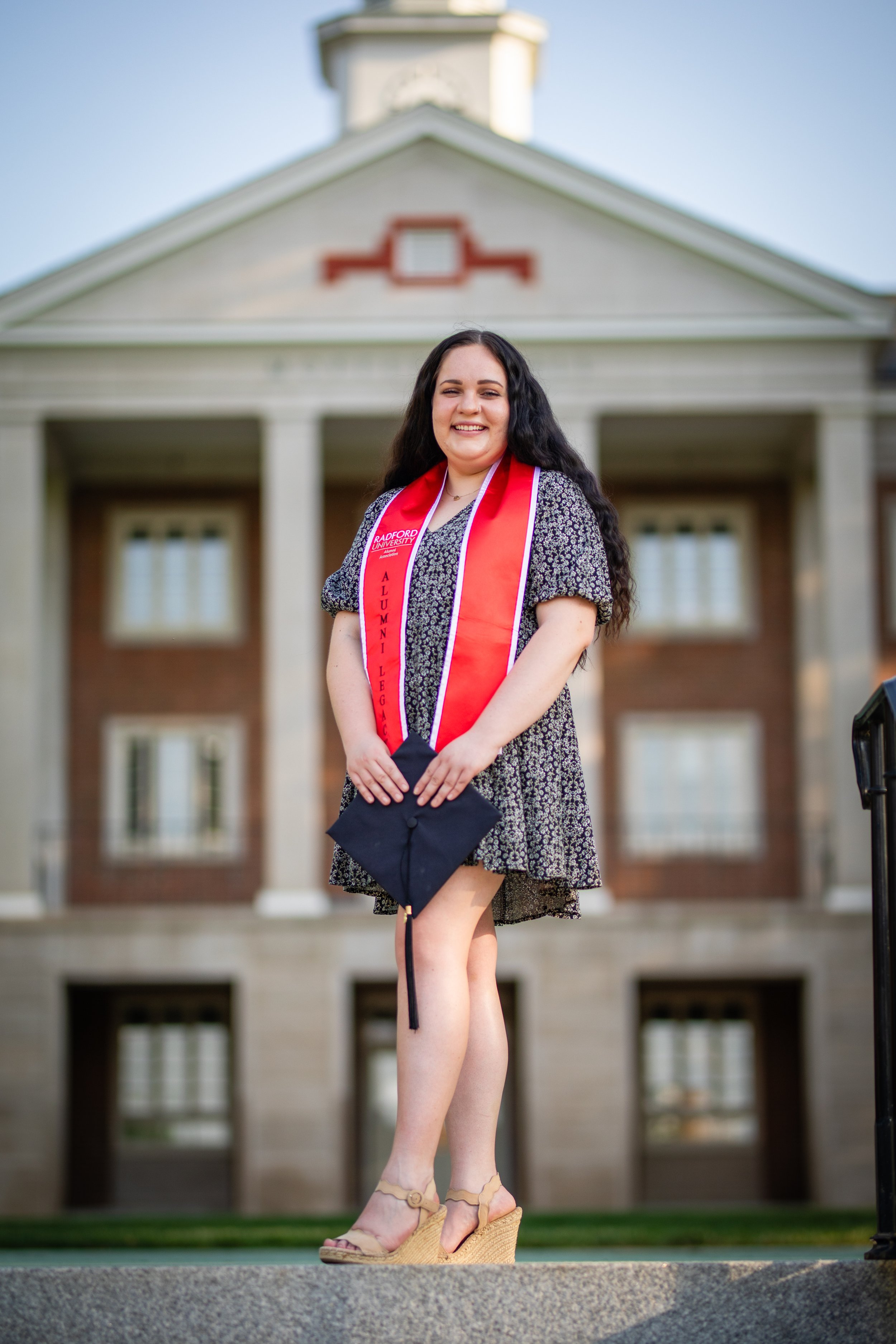 A young woman in a floral dress holding a graduation cap, standing in front of a large brick building with columns, celebrating her graduation with a red stole that reads 'Alumni' and 'Bradford University'.