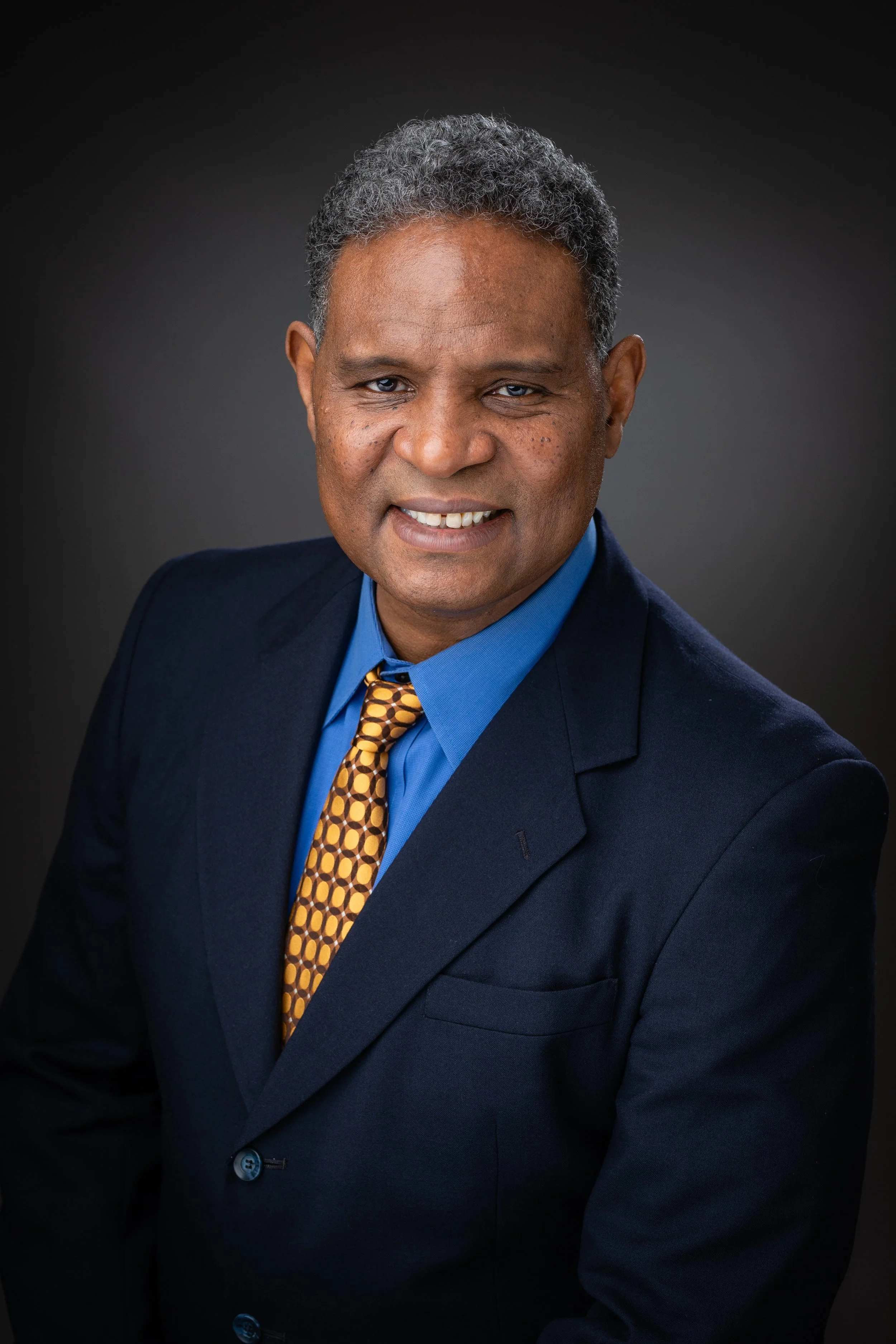 Professional portrait of a middle-aged man with gray hair, wearing a navy suit, blue shirt, and patterned yellow tie, smiling against a dark background.