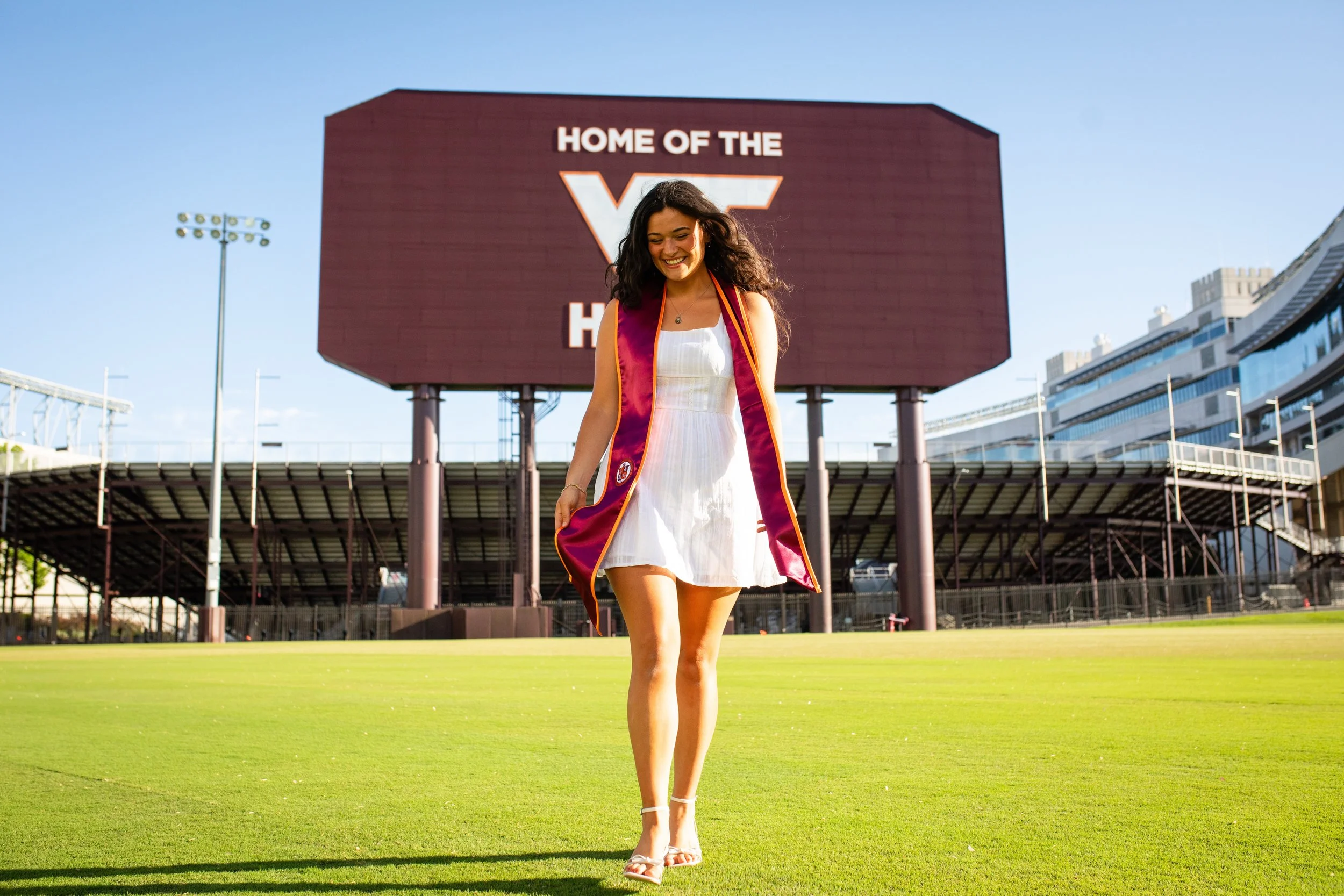 Young woman in a white dress and maroon graduation gown walking on a sports field in front of a large digital scoreboard that reads 'Home of the'.