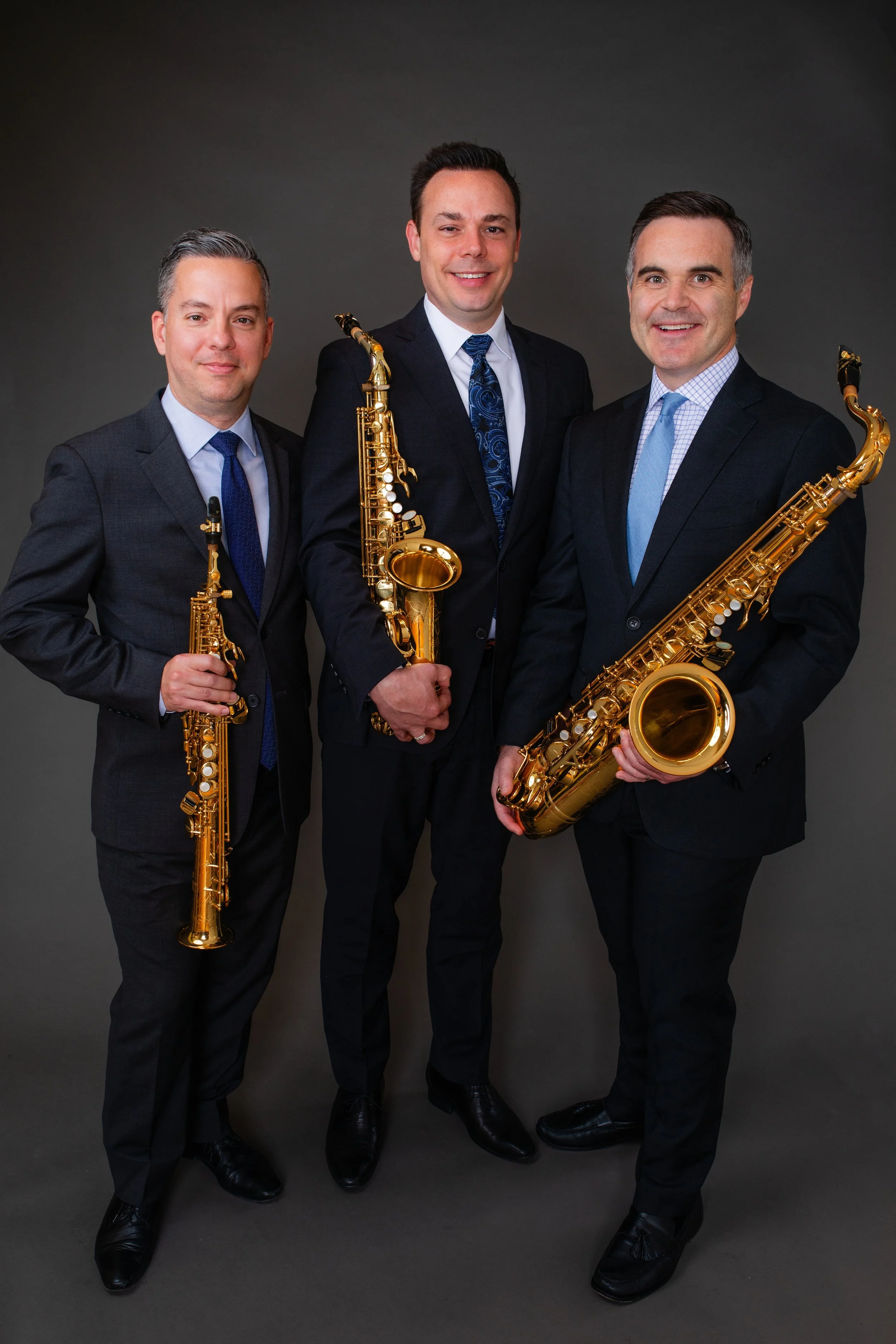 Three men dressed in suits holding saxophones in a professional photo studio.