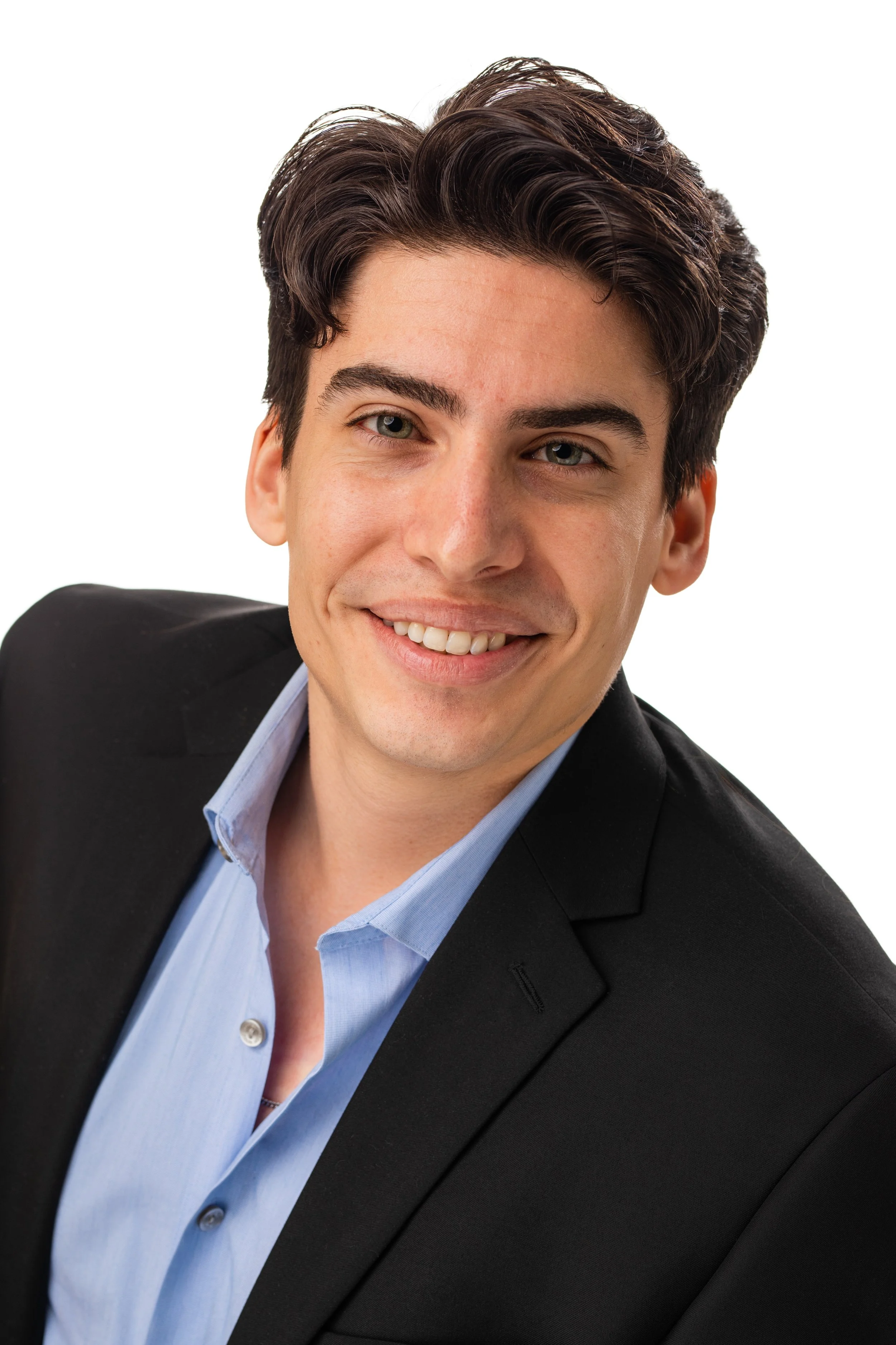 A young man with dark hair, light skin, wearing a black suit jacket and a light blue dress shirt, smiling with his head slightly tilted.