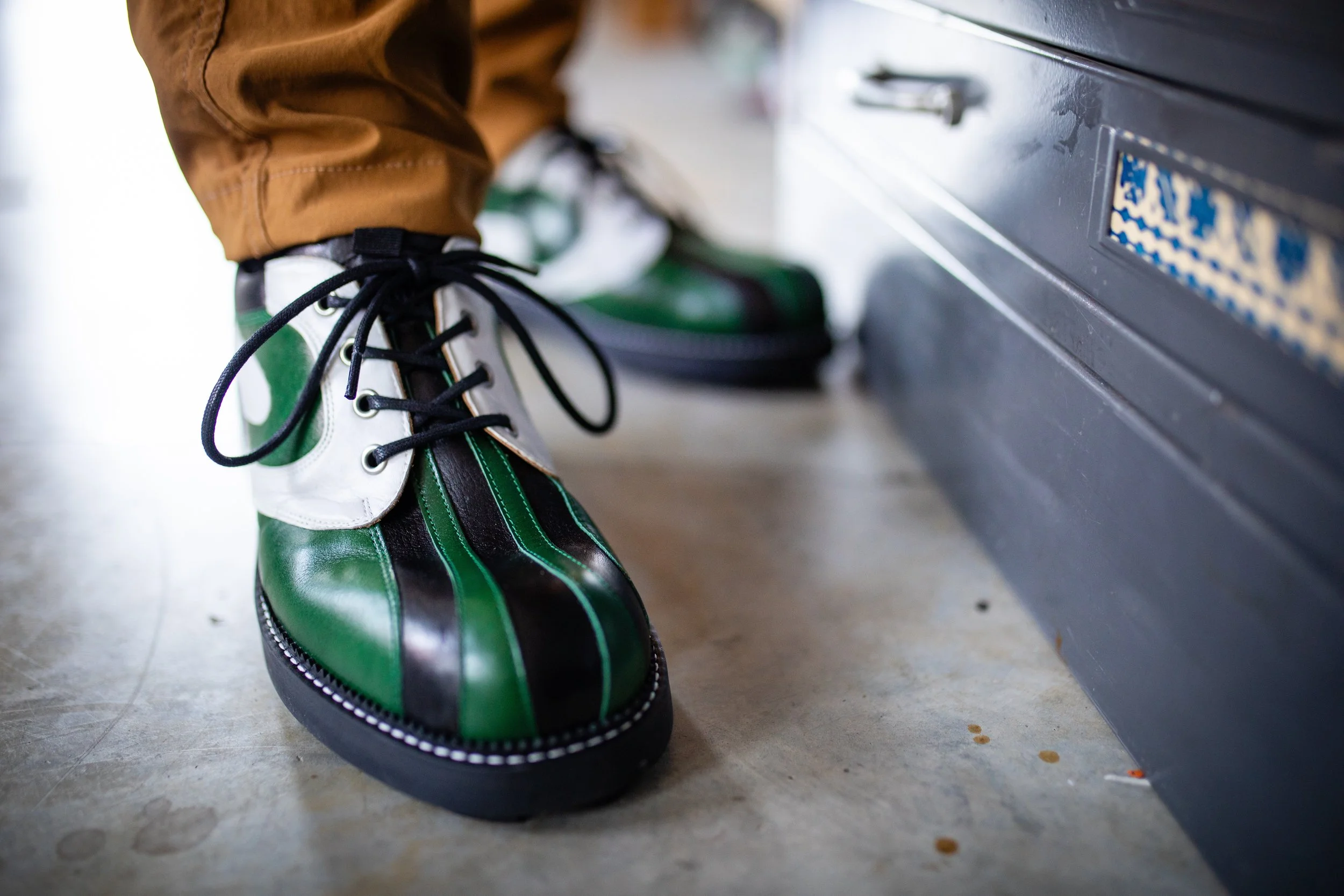 Close-up of a person wearing green, black, and white bowling shoes with black laces, stepping near a shoe locker or storage unit.