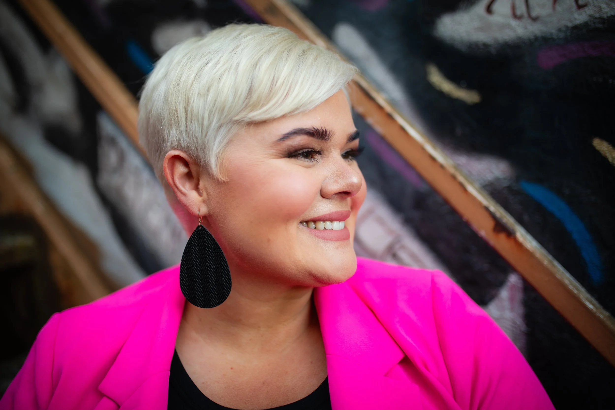 A woman with short blonde hair and a bright pink blazer, smiling and looking to her left, with a black earring and art in the background.