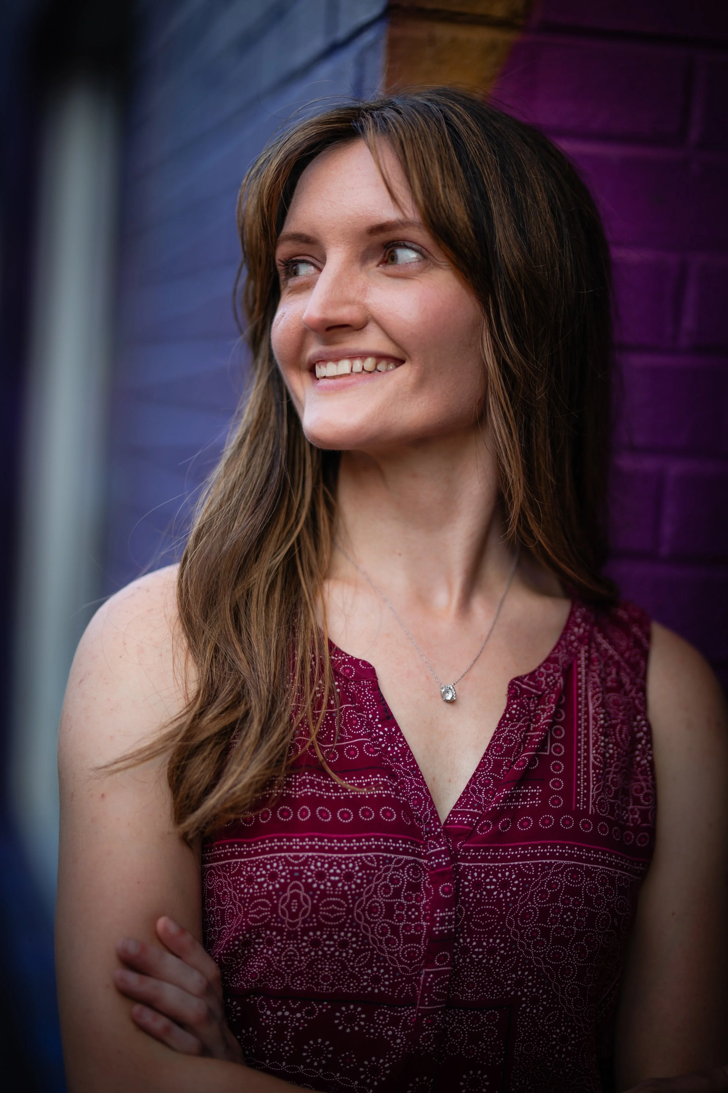 A young woman with brown hair and a smile, wearing a sleeveless maroon dress with white patterns, and a silver necklace, standing against a purple brick wall.