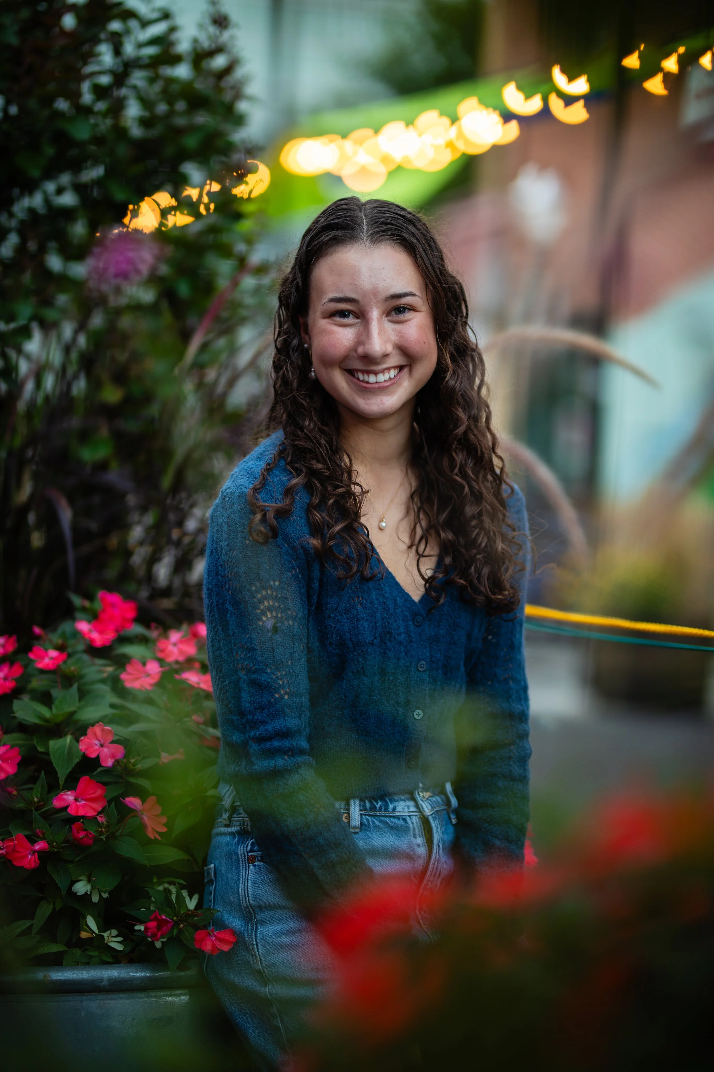 A young woman with long curly brown hair, wearing a blue knit sweater and jeans, smiling outdoors at dusk surrounded by pink and red flowers and greenery, with blurred warm string lights in the background.