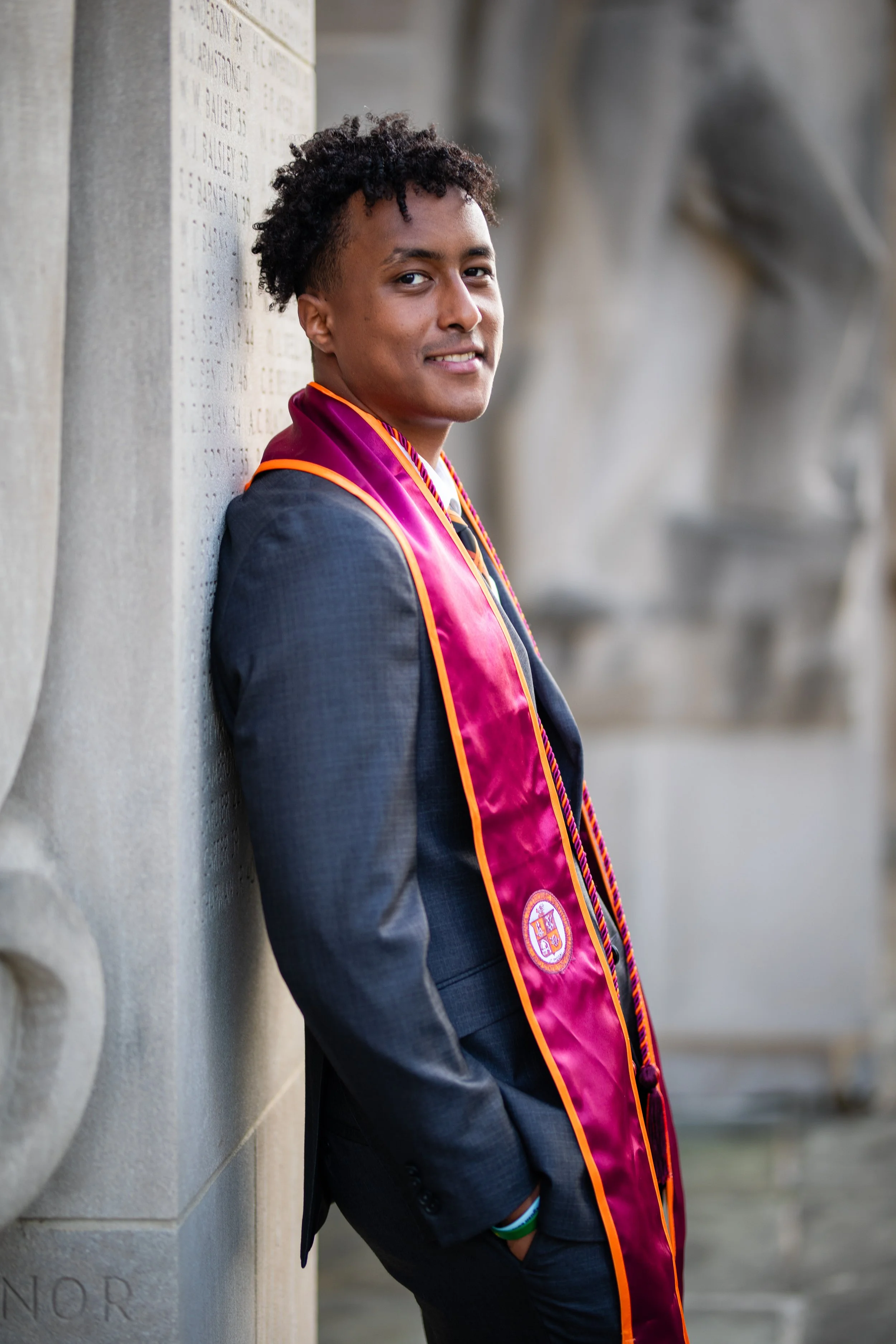 A young man in formal attire with a maroon and orange graduation stole standing against a stone wall.