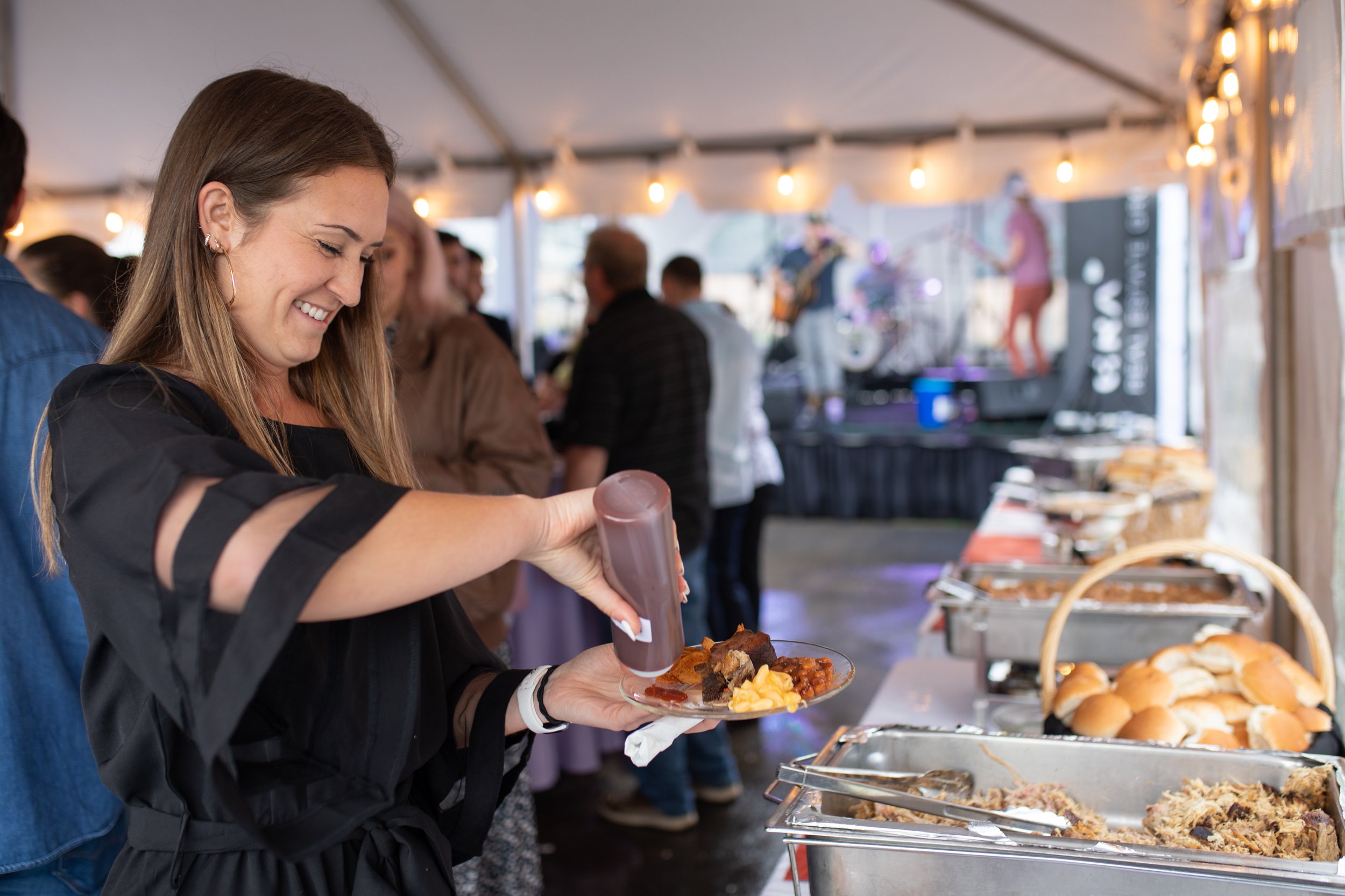 Smiling woman serving herself barbecue plate at buffet table with buns and mac and cheese, in tent with string lights and live music stage in background.