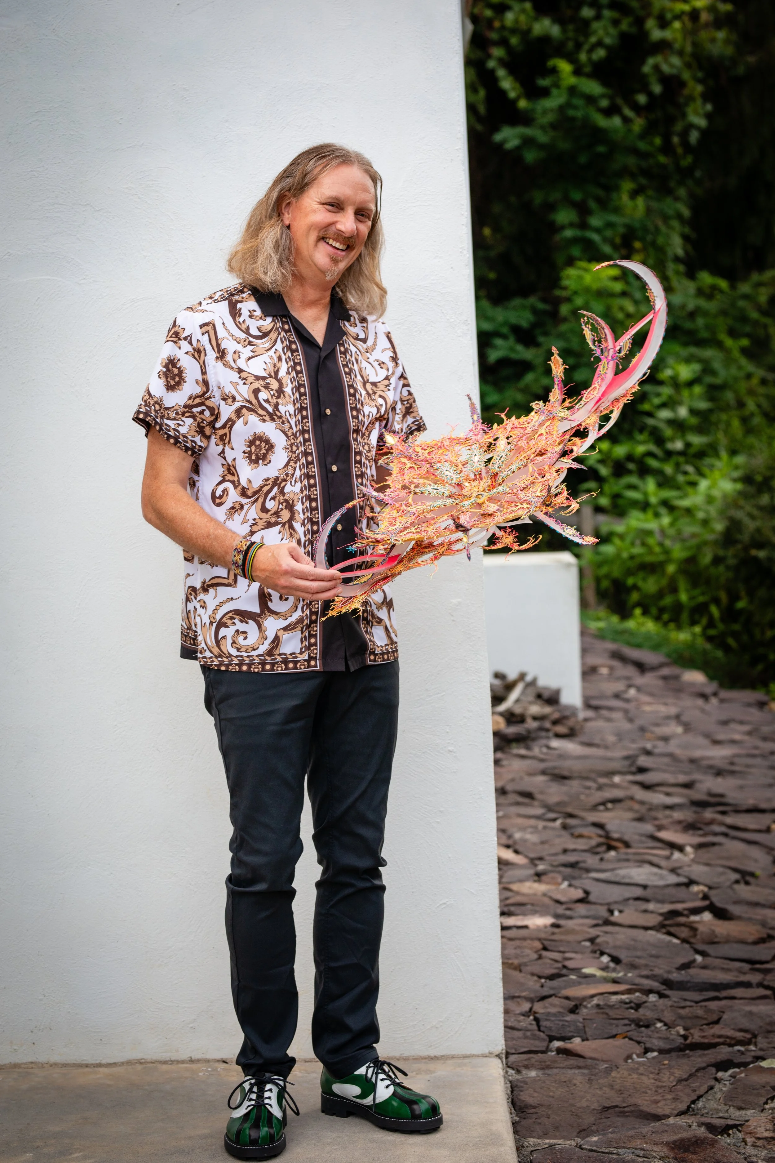 A man with long hair, smiling, standing outdoors against a white wall, holding a colorful, elaborate glass sculpture resembling a flower or sea creature, with greenery in the background.