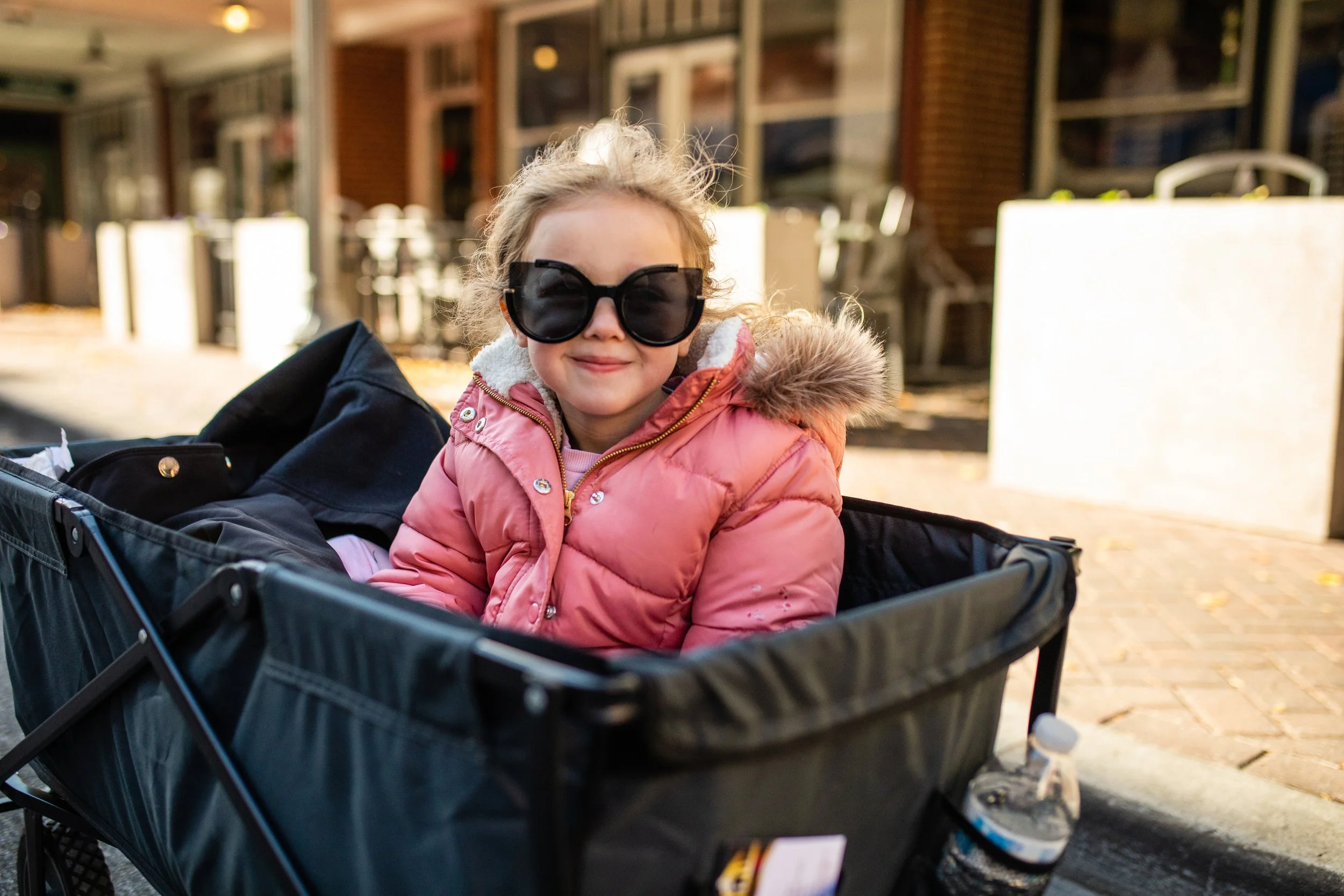 Young girl wearing large sunglasses and a pink jacket sitting in a shopping cart outdoors on a sunny day