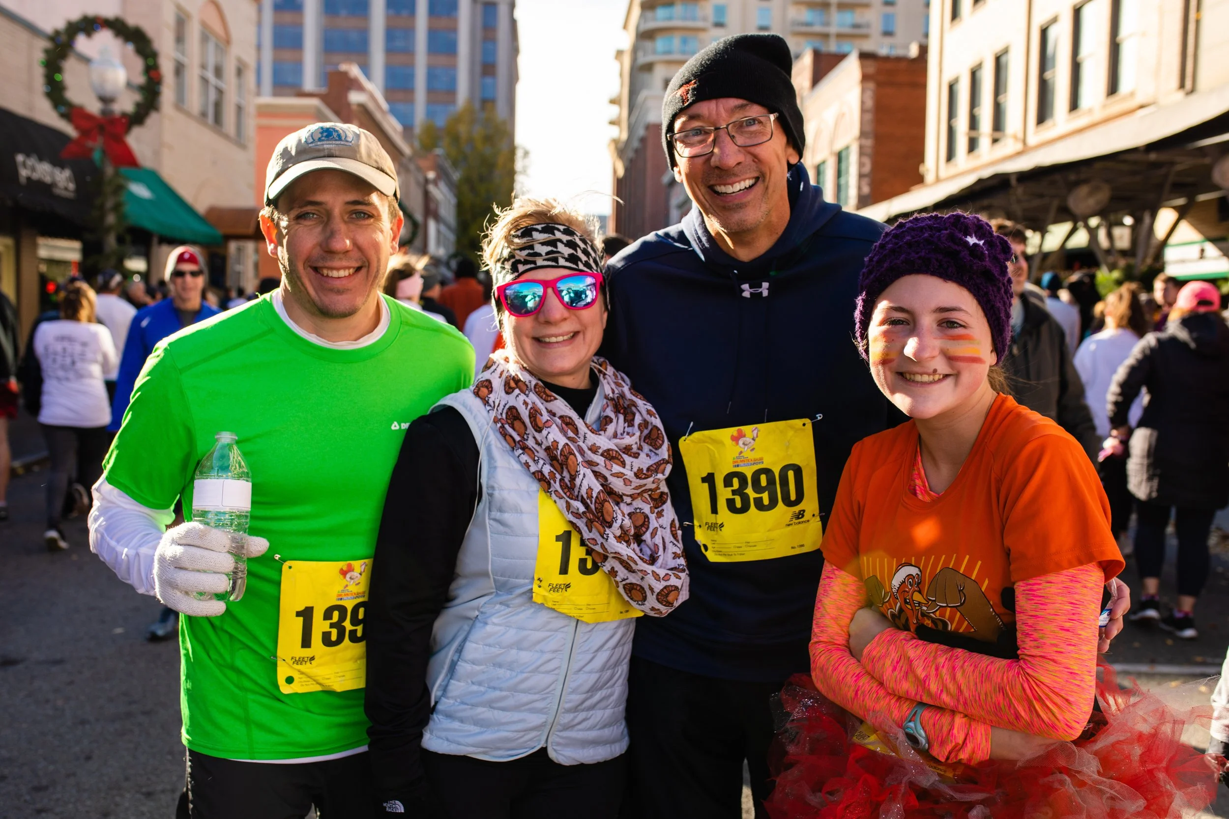 A group of four people smiling at a race event, with bib numbers 1390 and 1393, surrounded by other participants on a city street.