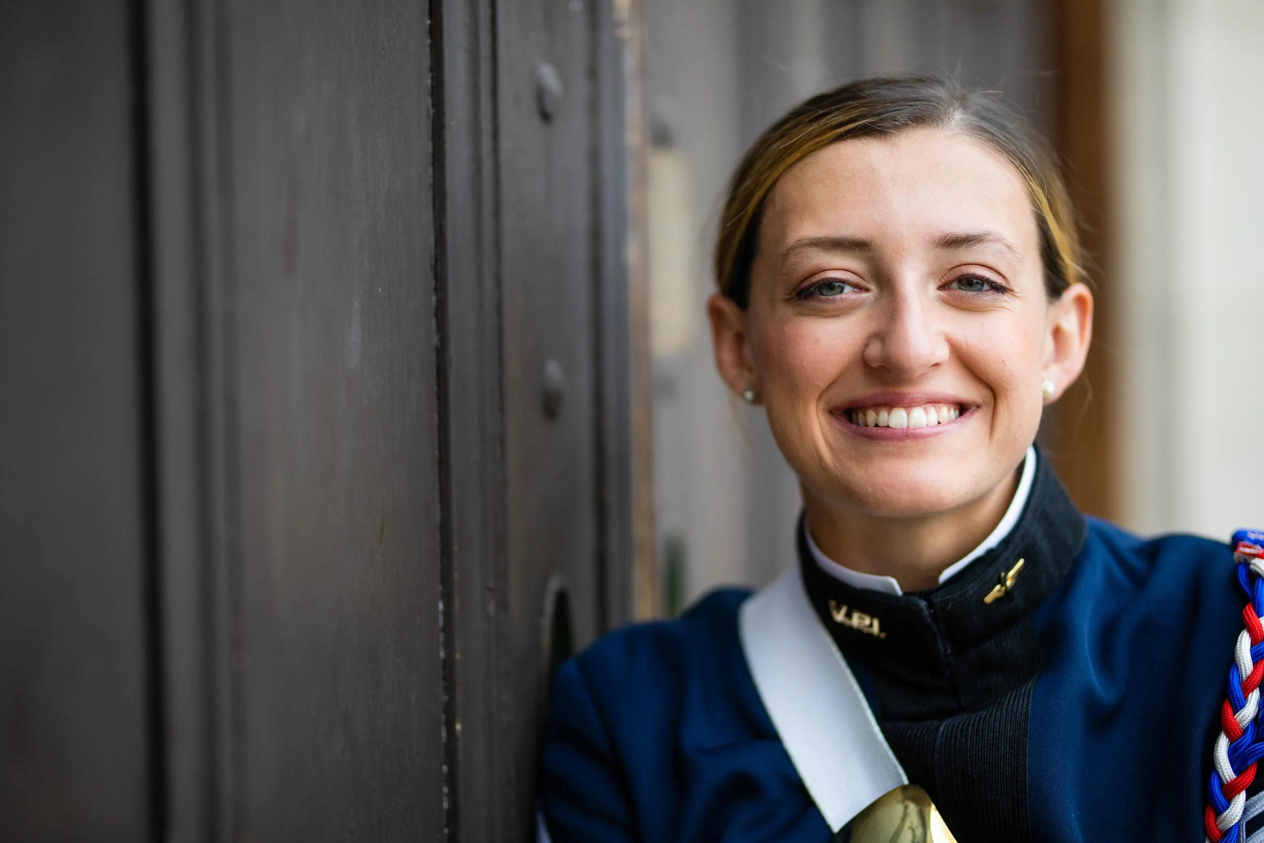 Close-up of a smiling woman in a dark blue and white choir uniform, leaning against a wooden wall.
