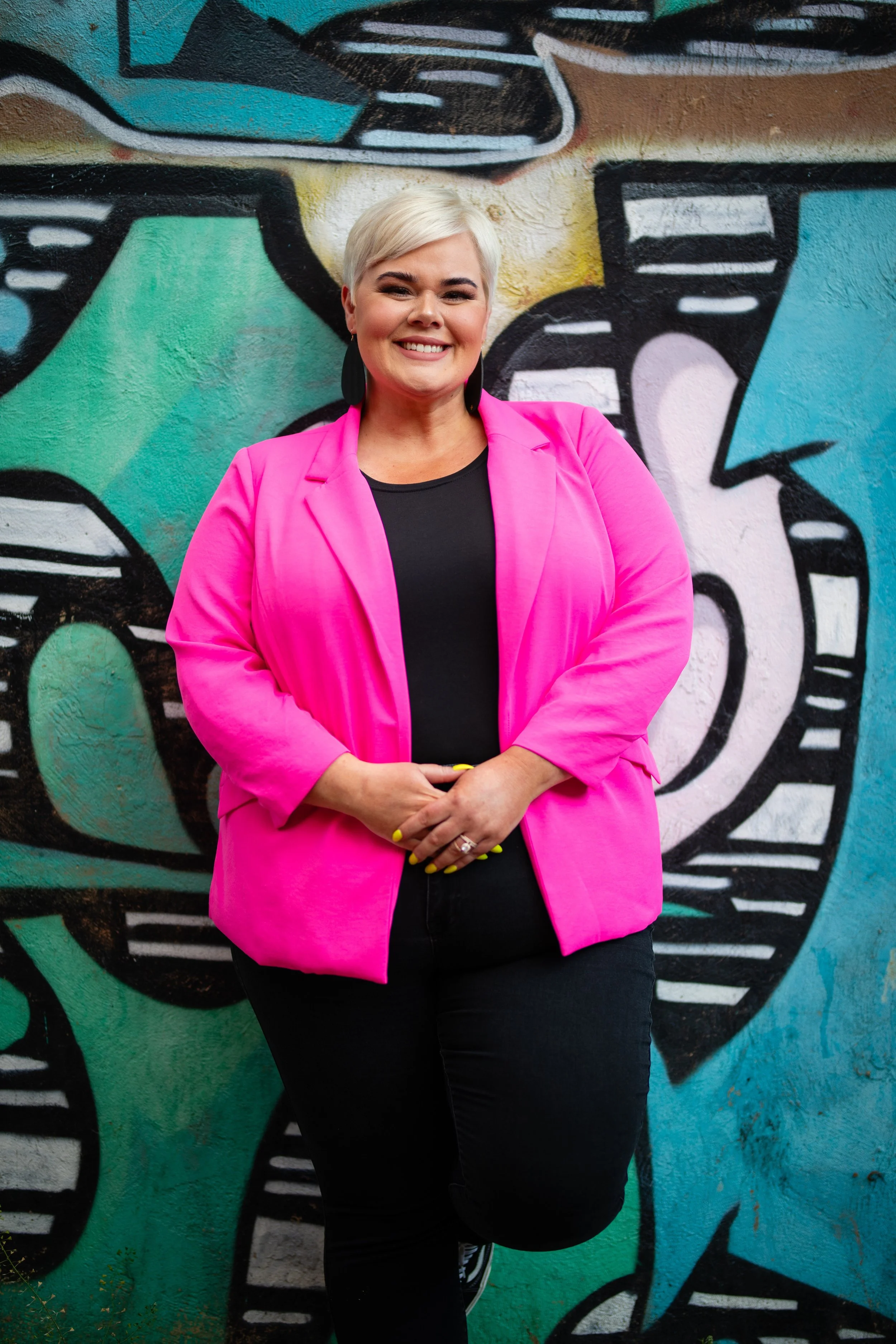 A woman with short blonde hair smiling while standing in front of a colorful graffiti mural. She is wearing a bright pink blazer over a black top, with black pants and yellow nails.