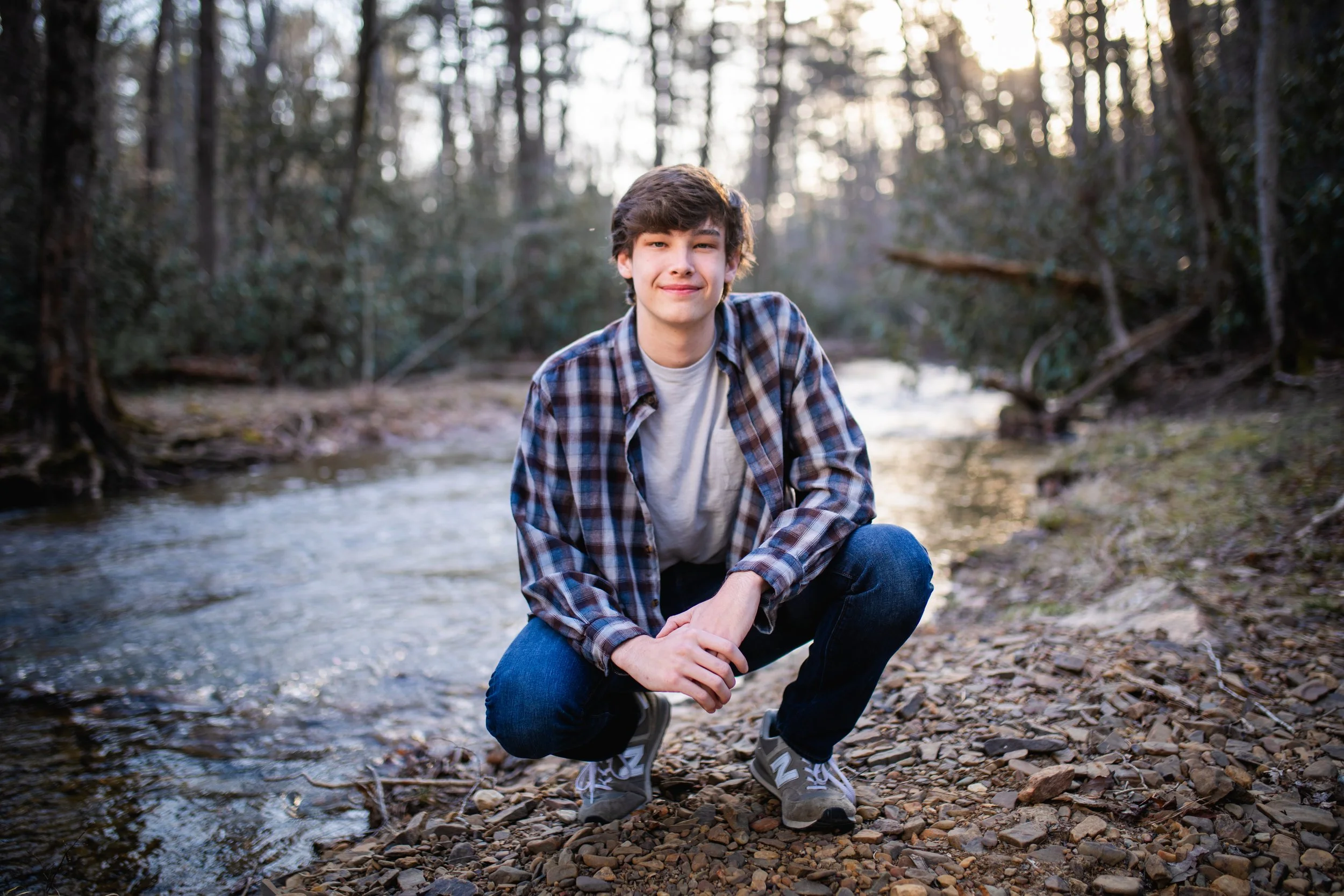 Young boy squatting on rocky riverbank with trees in background during sunset.
