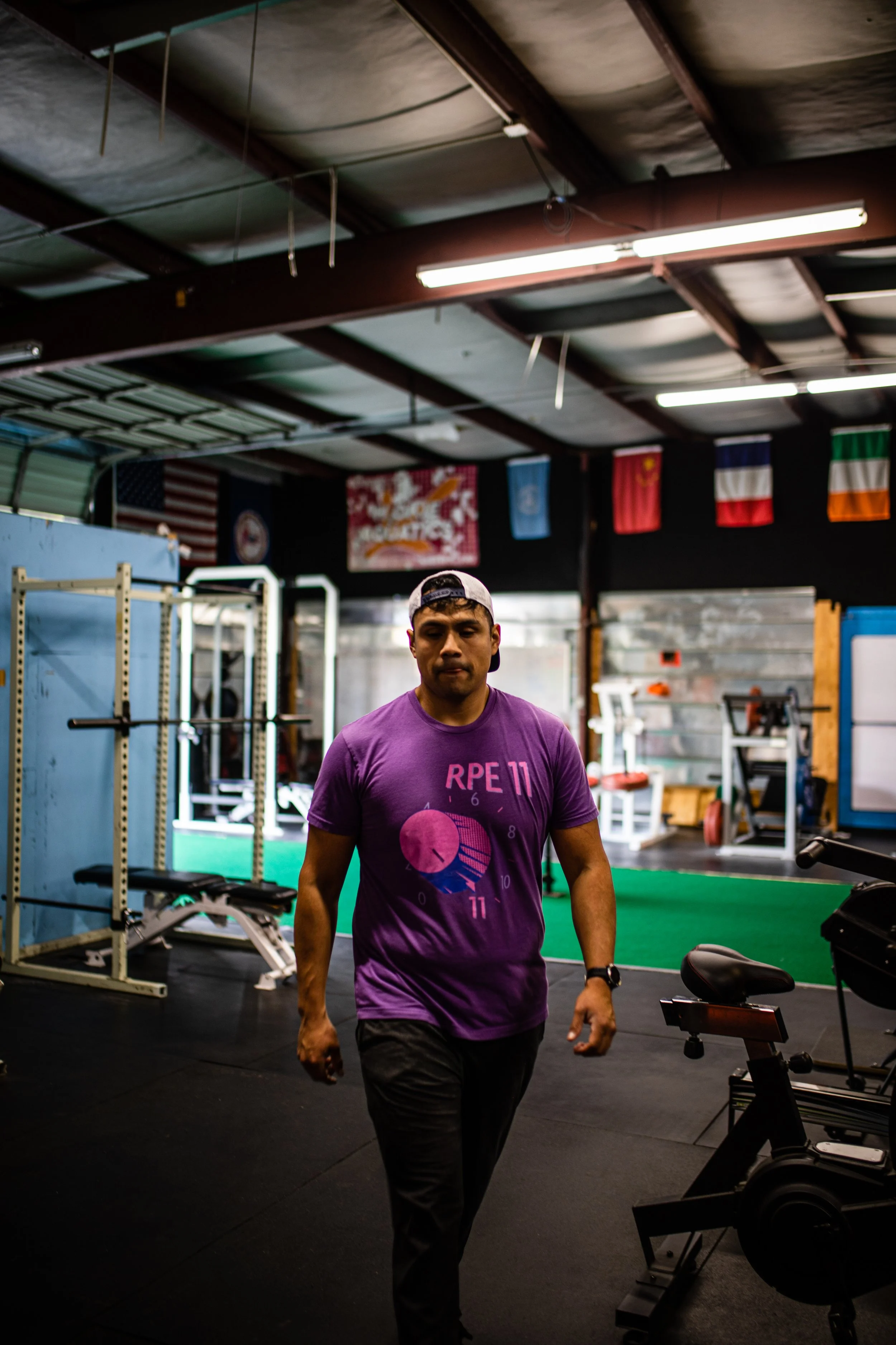 Man walking in a gym, wearing a purple T-shirt and a white cap backward, with workout equipment in the background.
