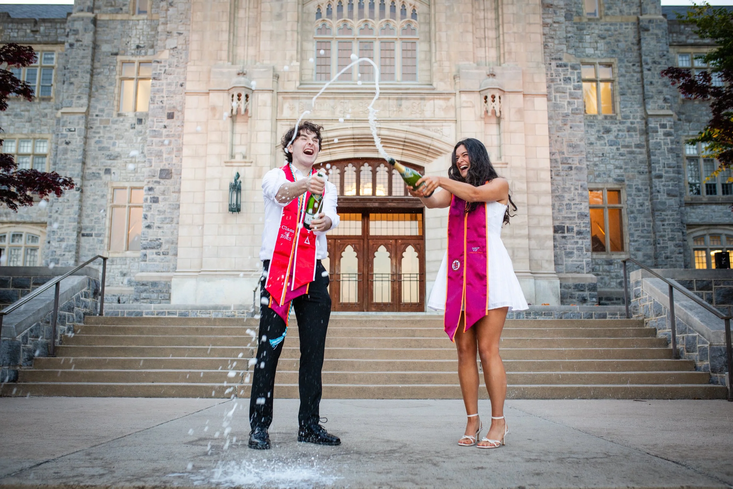 Two graduates celebrating on the steps of a university, cheering and spraying champagne.