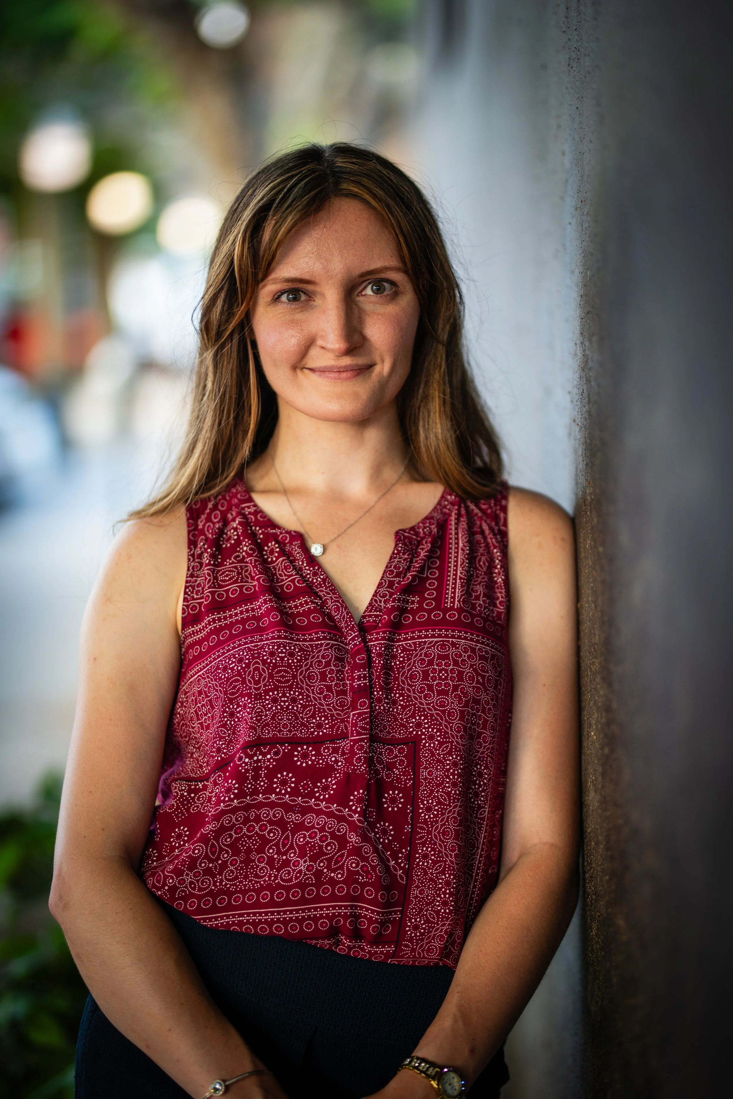A woman with long brown hair, wearing a sleeveless red patterned blouse, standing outdoors against a gray wall, with blurred trees and park in the background.