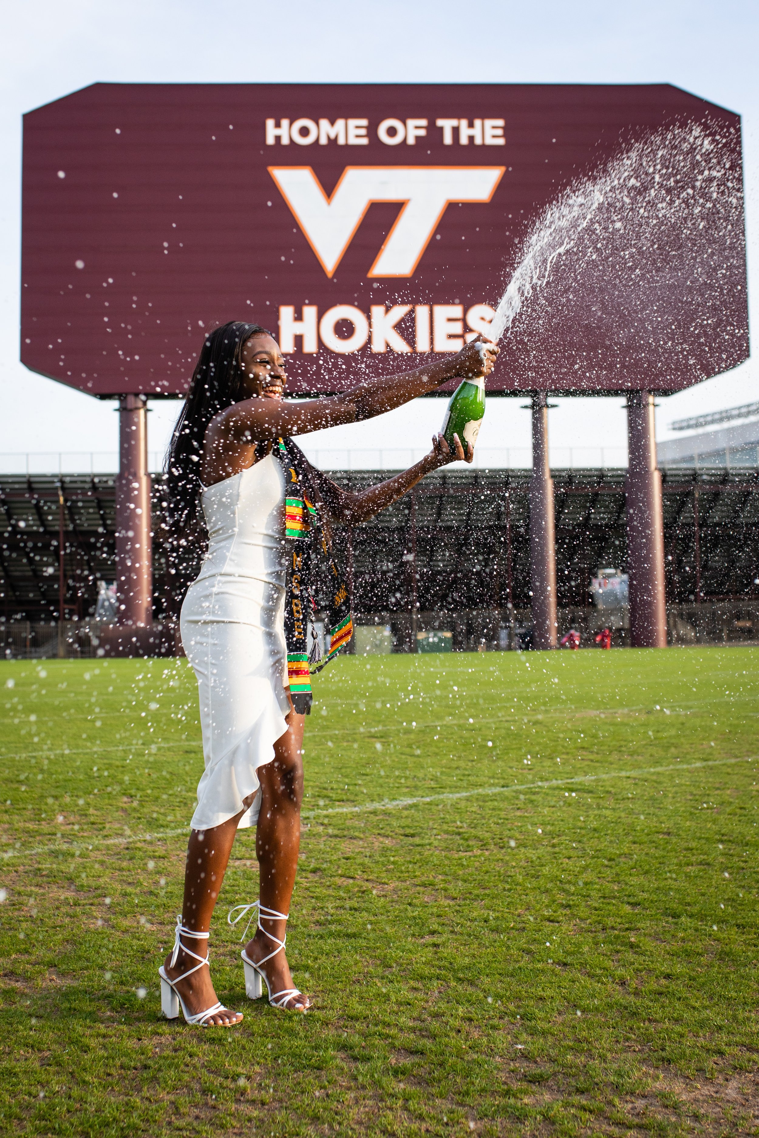 Woman celebrating with champagne on a football field, in front of a sign that reads 'Home of the VT Hokies'.