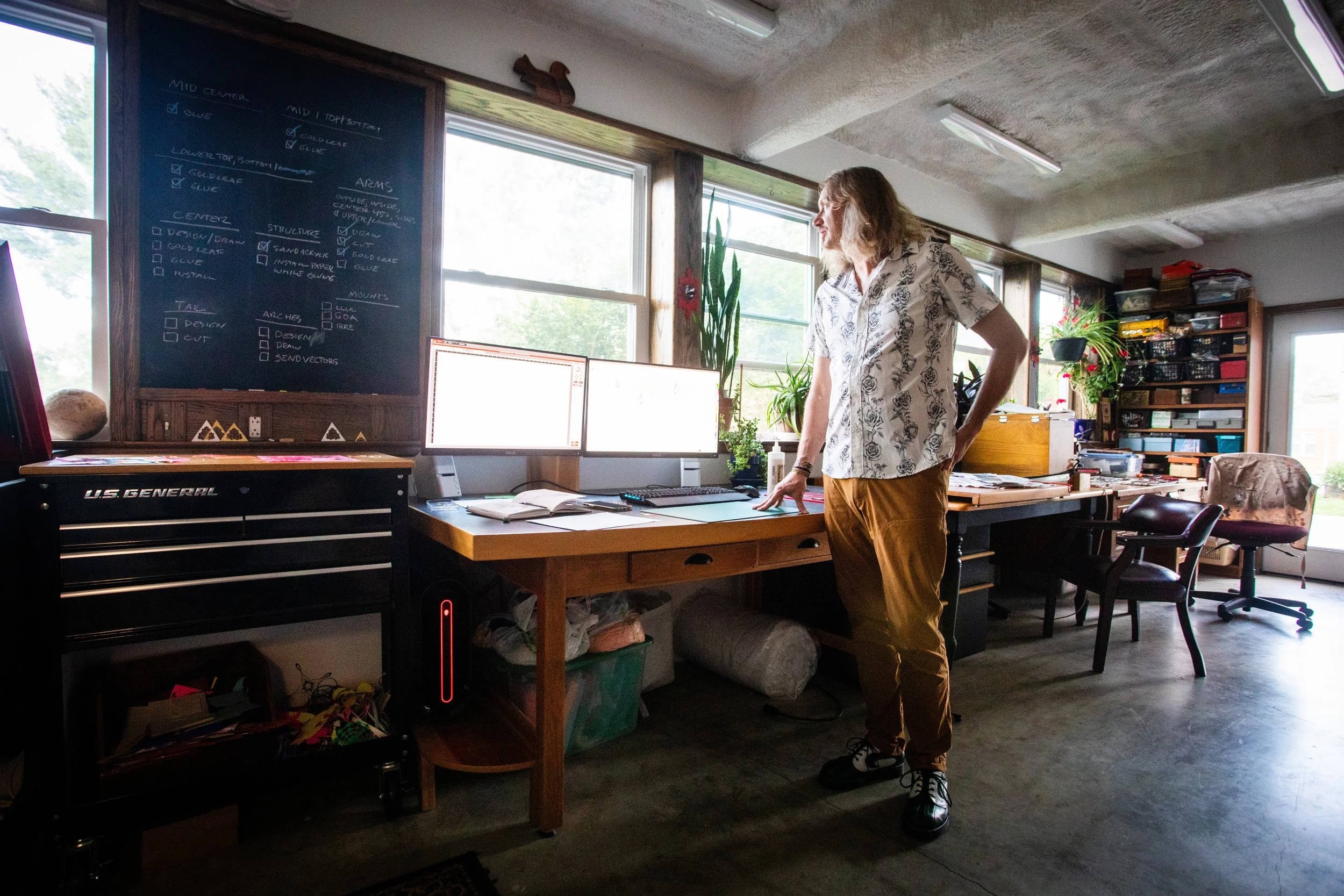 A person in a patterned shirt and brown pants standing in an office or workshop space, looking at a desk with dual computer monitors, books, and various office supplies. There is a chalkboard with notes on the wall behind.