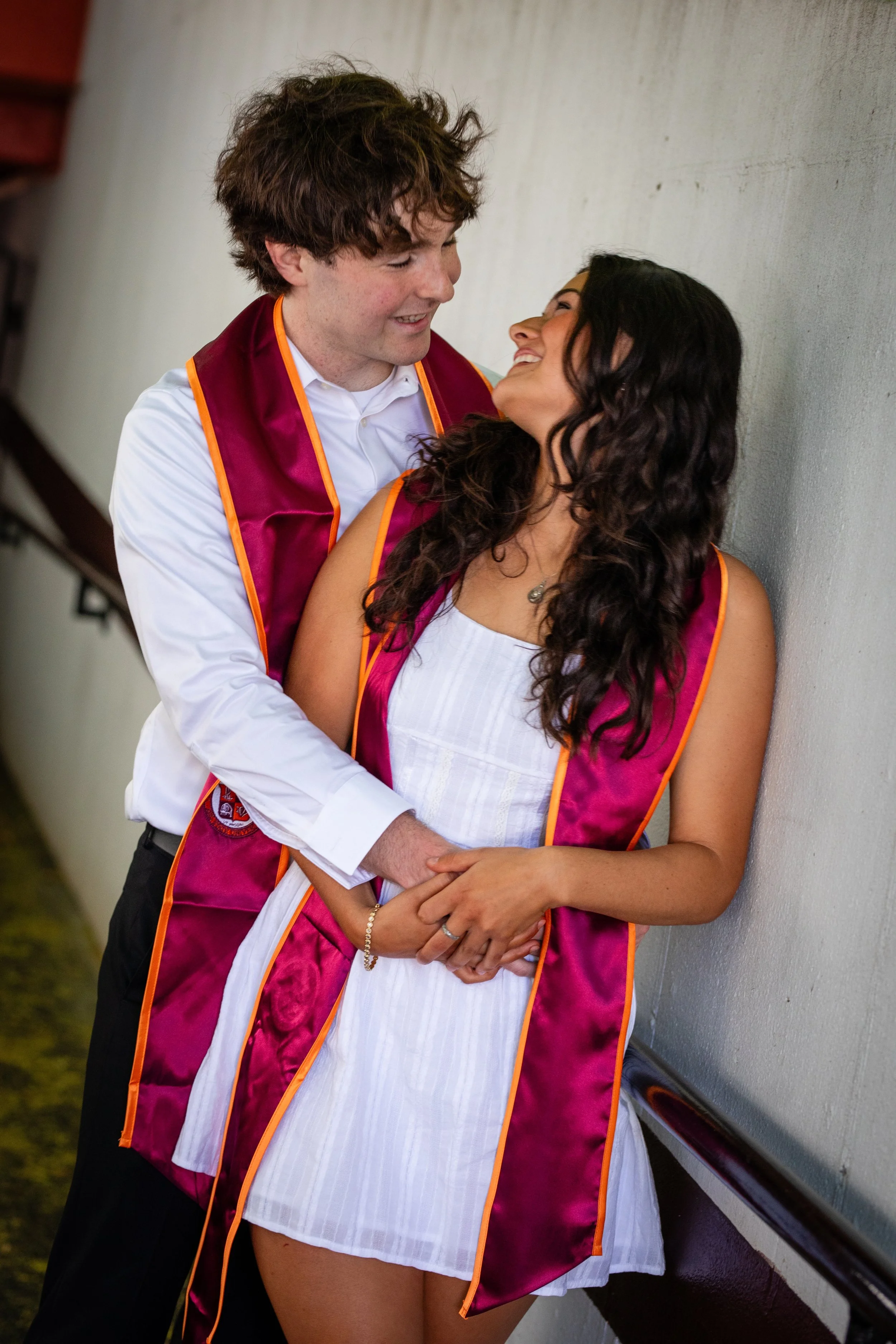 A young man and woman in graduation caps and gowns smiling and looking at each other, standing against a wall.
