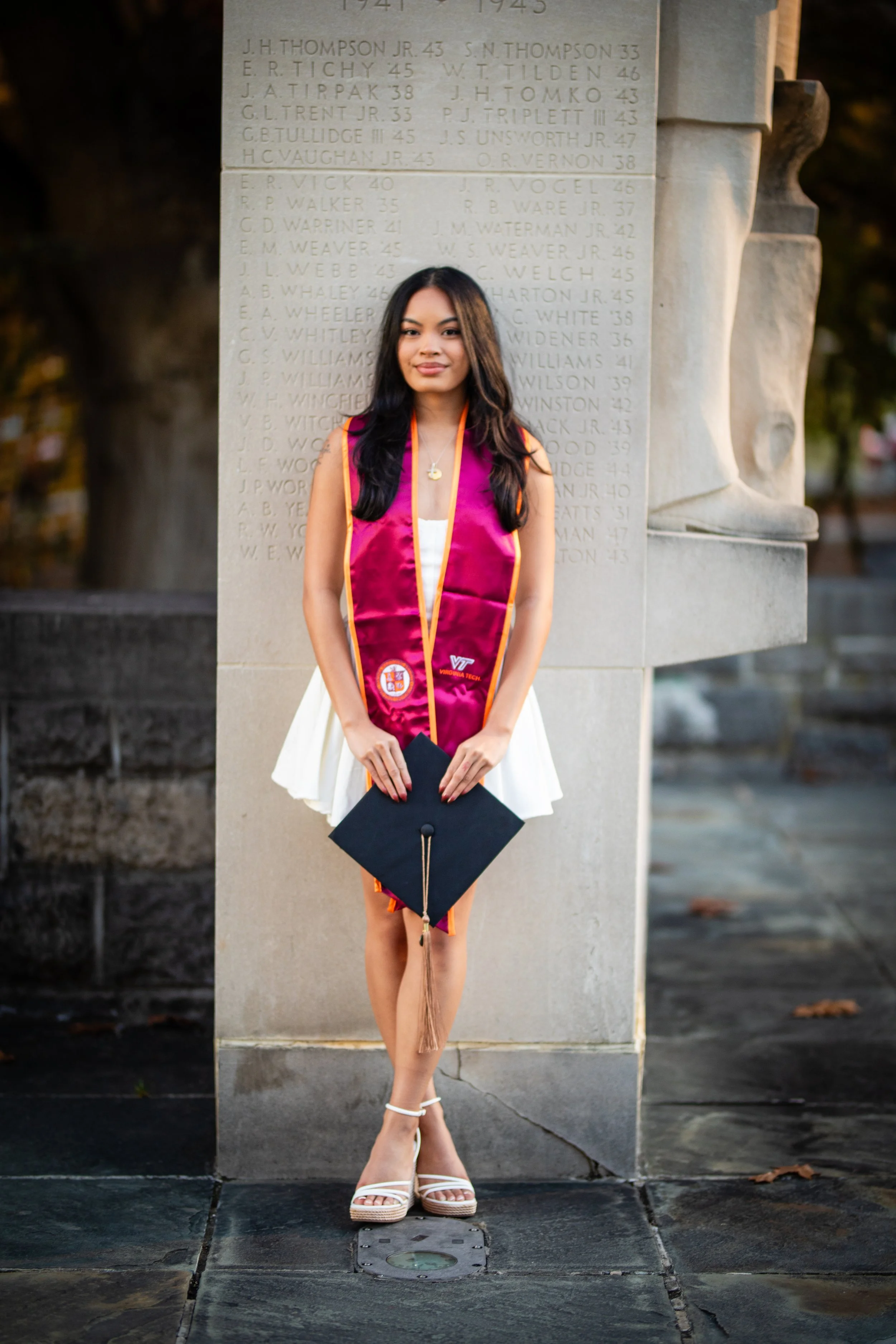 A young woman in a white dress and white heels standing in front of a monument, wearing a maroon graduation stole, holding a graduation cap, with a stone wall with engraved names behind her.