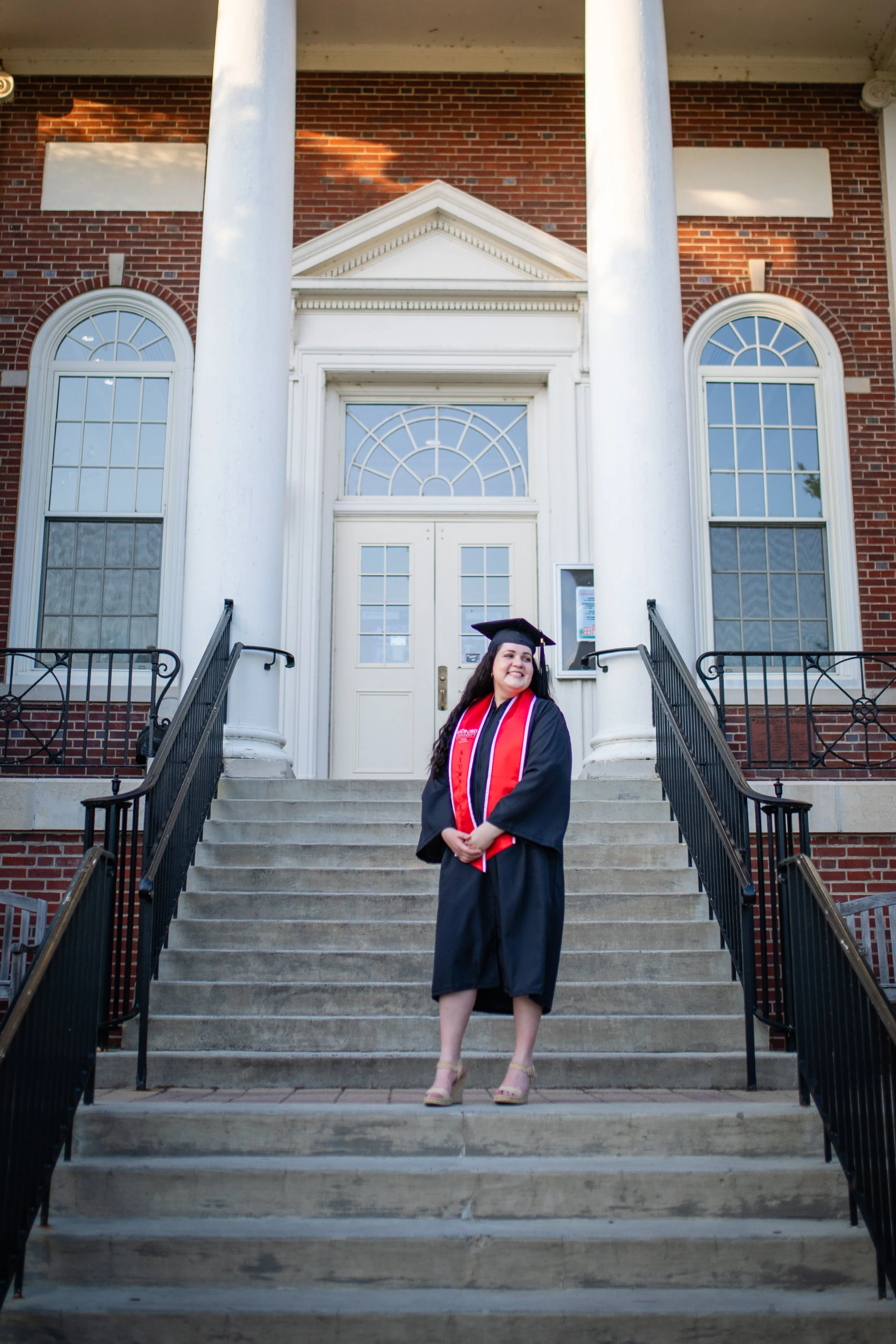 A woman in a graduation cap and gown standing on the steps of a brick building with white columns.
