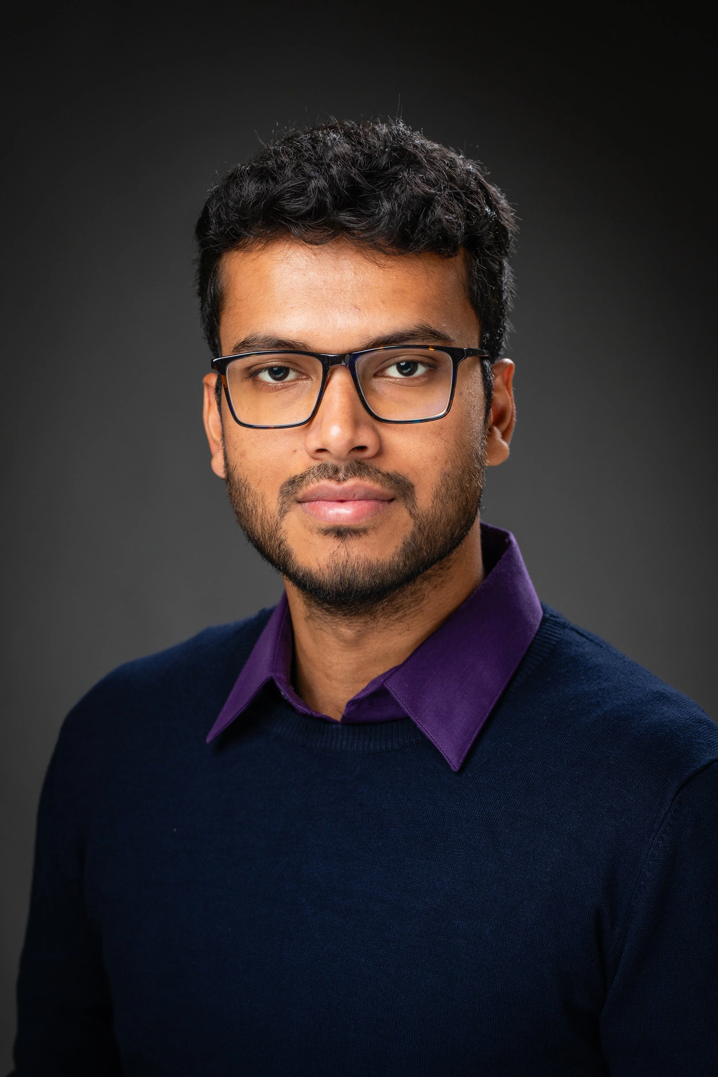 A man with glasses, dark hair, and a beard, wearing a dark sweater over a purple collared shirt, against a dark gray background.