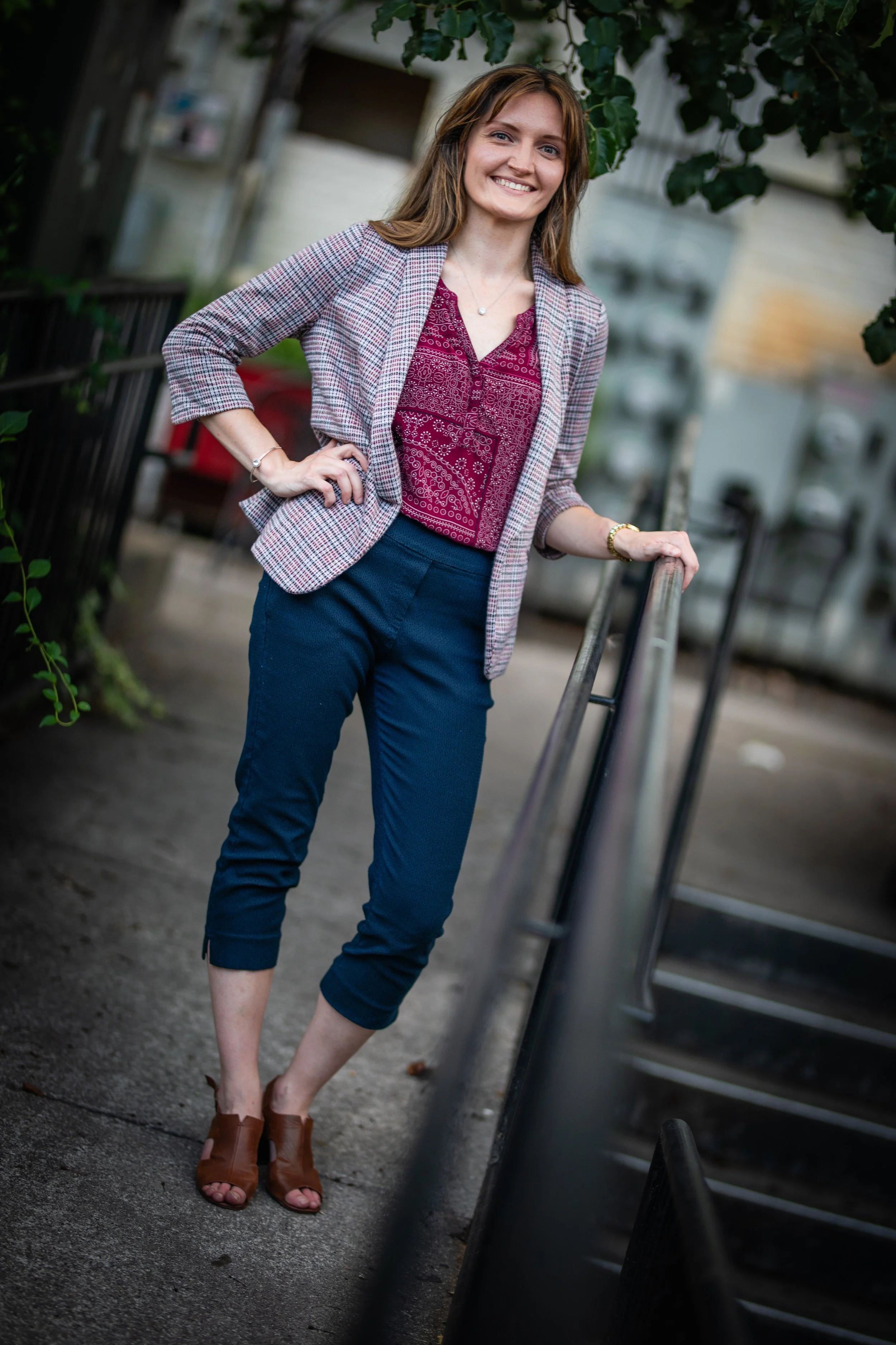 A woman standing outdoors on a sidewalk, smiling, wearing a plaid blazer, burgundy blouse, dark blue pants, and brown heeled sandals.