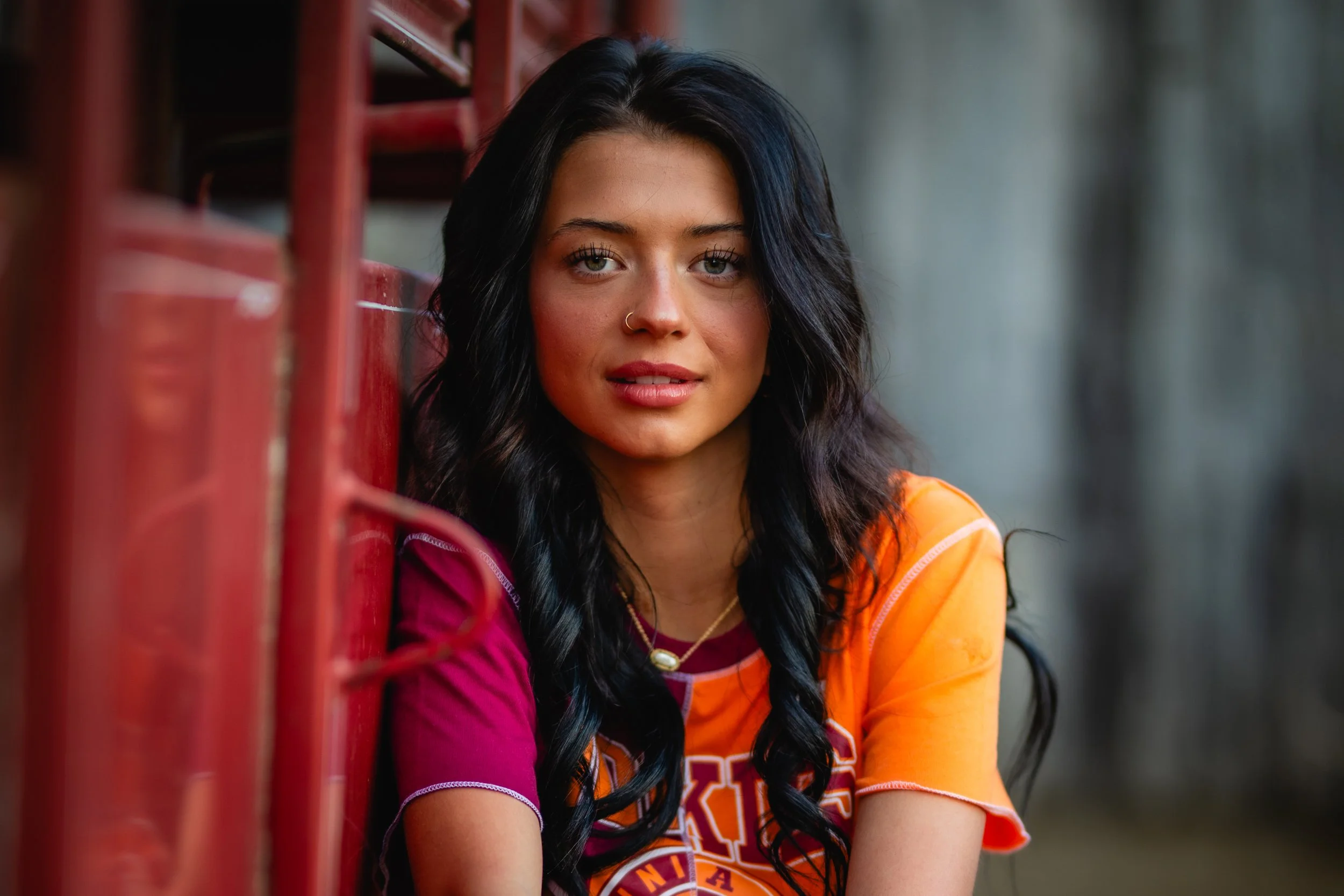 A young woman with long black hair and a nose piercing, wearing an orange and maroon sports jersey, sitting outdoors near a red metal structure, with a blurred natural background.